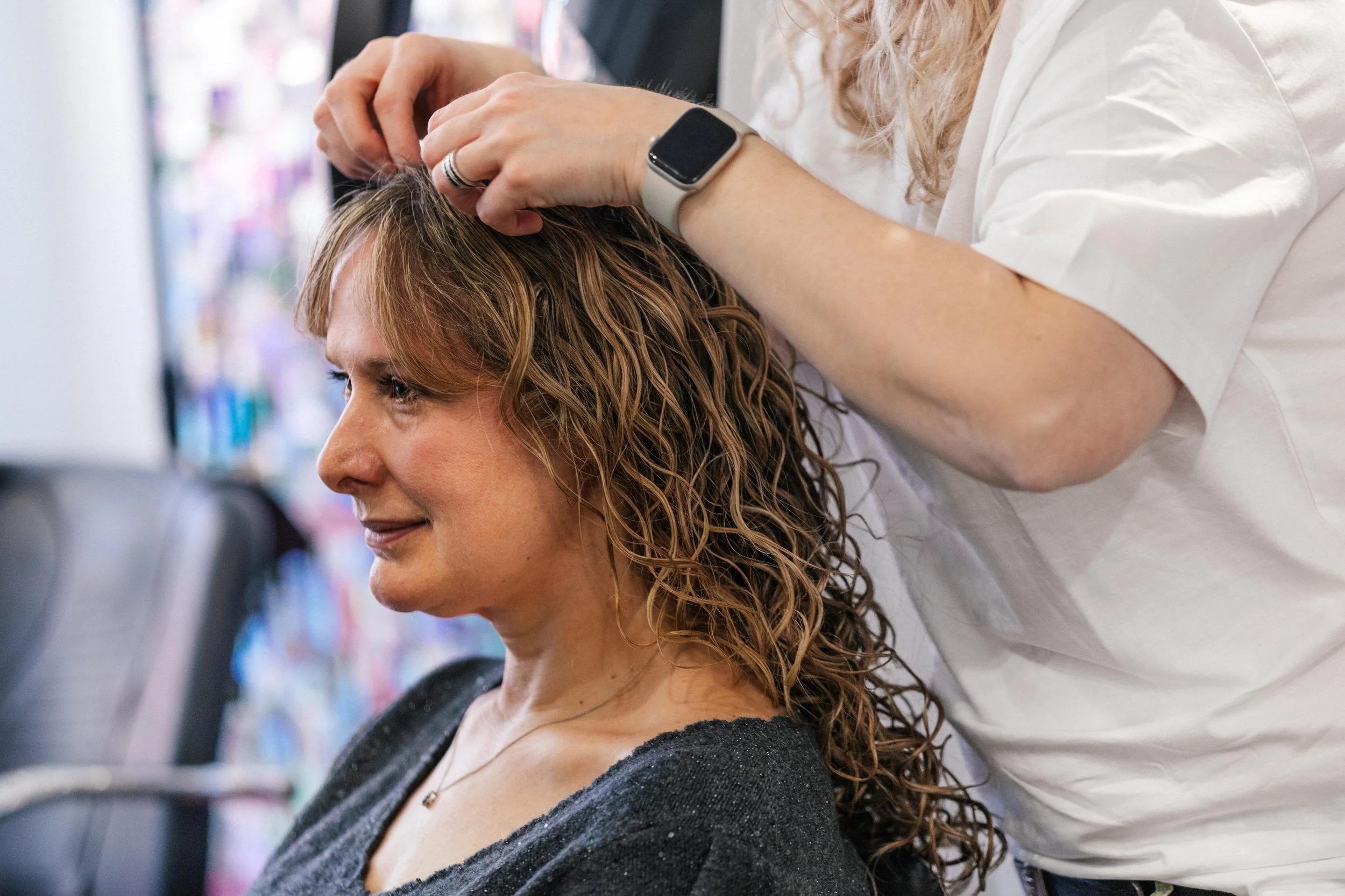 A woman with curly hair getting a haircut at a salon while sitting in a chair, with a hairstylist attending to her.