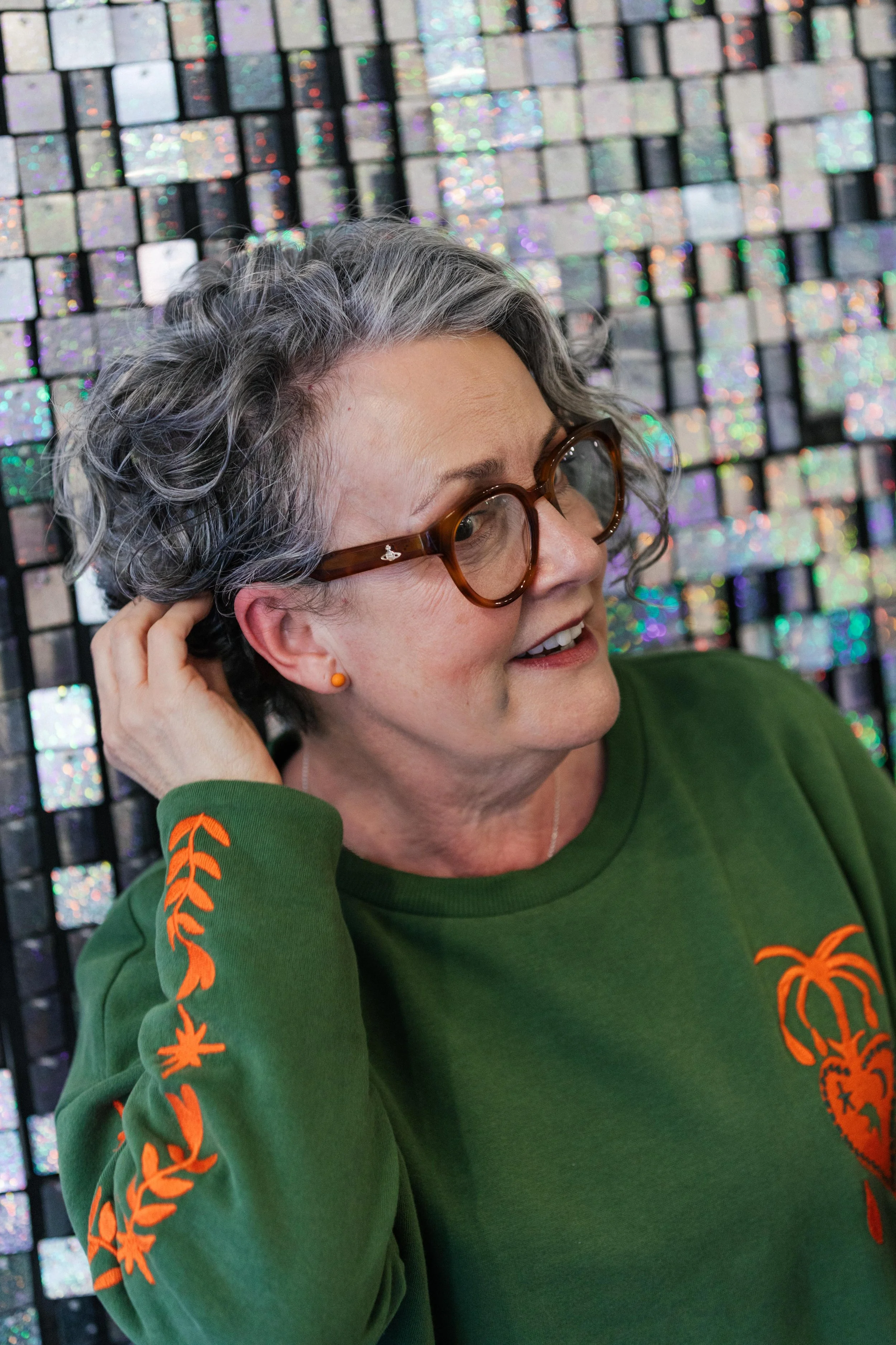 A woman with gray, curly hair, wearing glasses and a green sweatshirt with orange embroidery, pose against a holographic, tiled background.