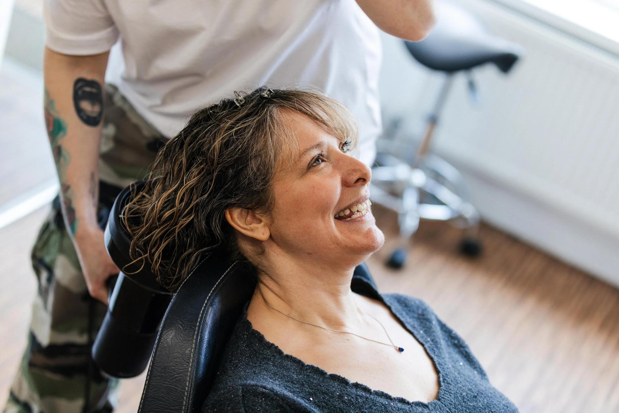 A woman with curly hair smiling while sitting in a salon chair, getting her hair washed or styled by a cosmetologist.