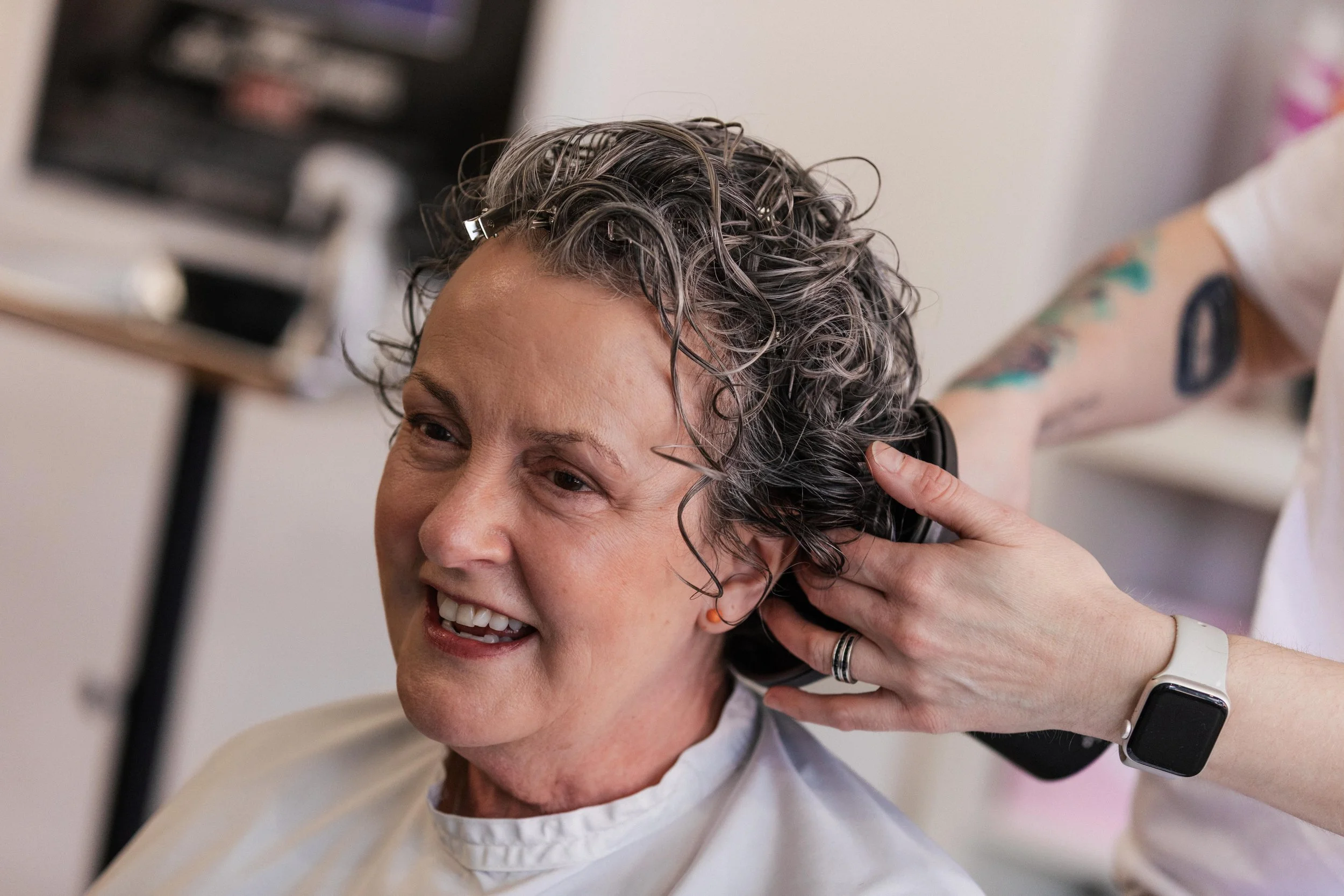 A woman with short, curly, gray hair is sitting in a chair while a hairdresser trims her hair. The woman is smiling and wearing a white salon cape. The hairdresser has colorful tattoos on her arm and is wearing a white wristwatch.