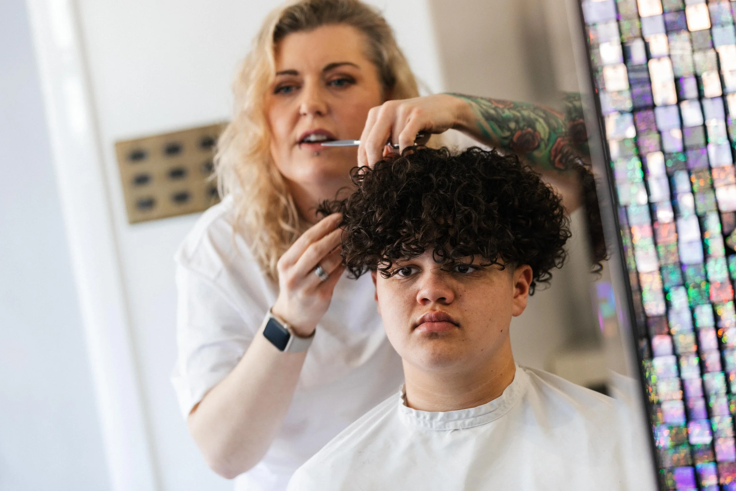 A woman with blonde hair getting her curly hair cut or styled at a salon, seated in front of a mirror displaying colorful reflections.