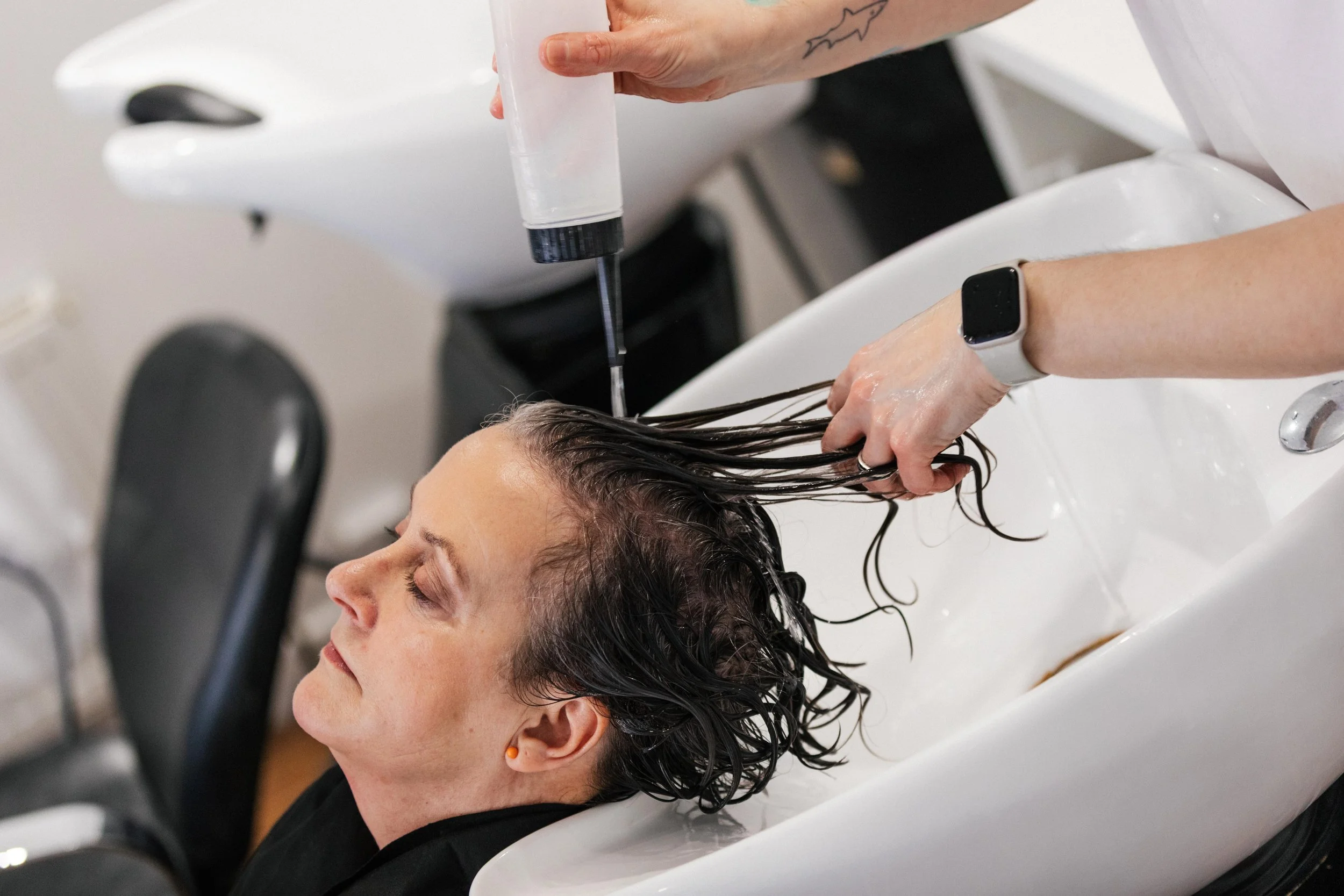 A woman reclining at a salon sink with her head in a basin, getting her hair washed by a stylist holding a shampoo bottle and rinsing her hair.