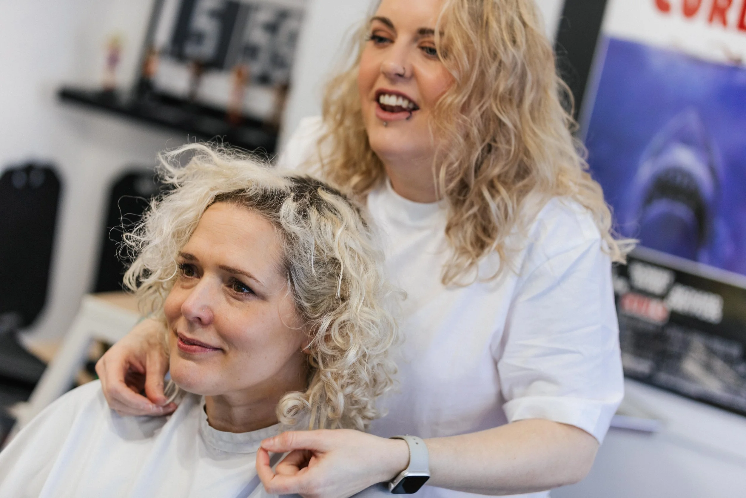 A hairstylist with curly blonde hair shows a woman with similar hair her reflection in a mirror at a salon.