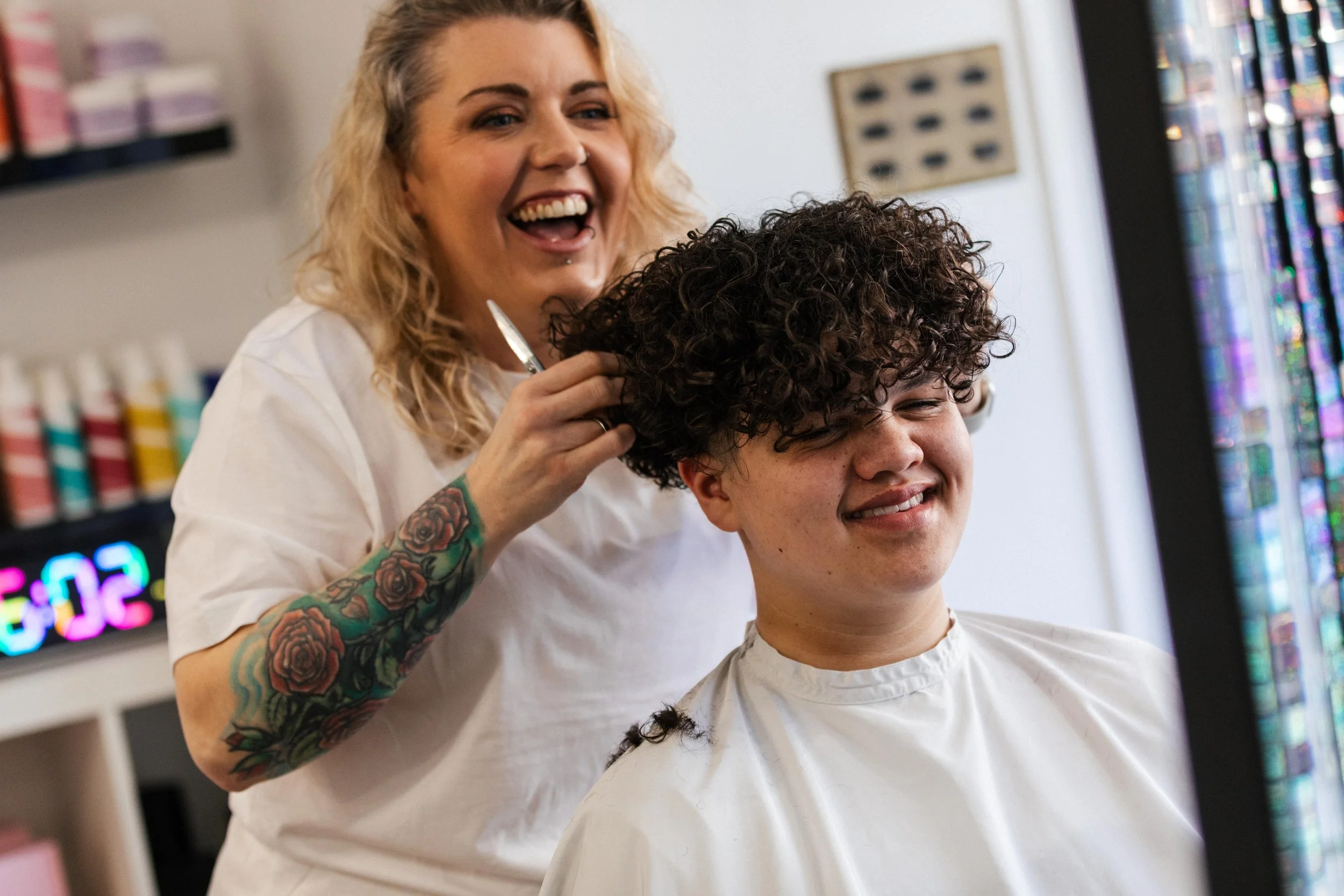 A hairstylist with tattoos trimming a young man's curly hair in a salon.