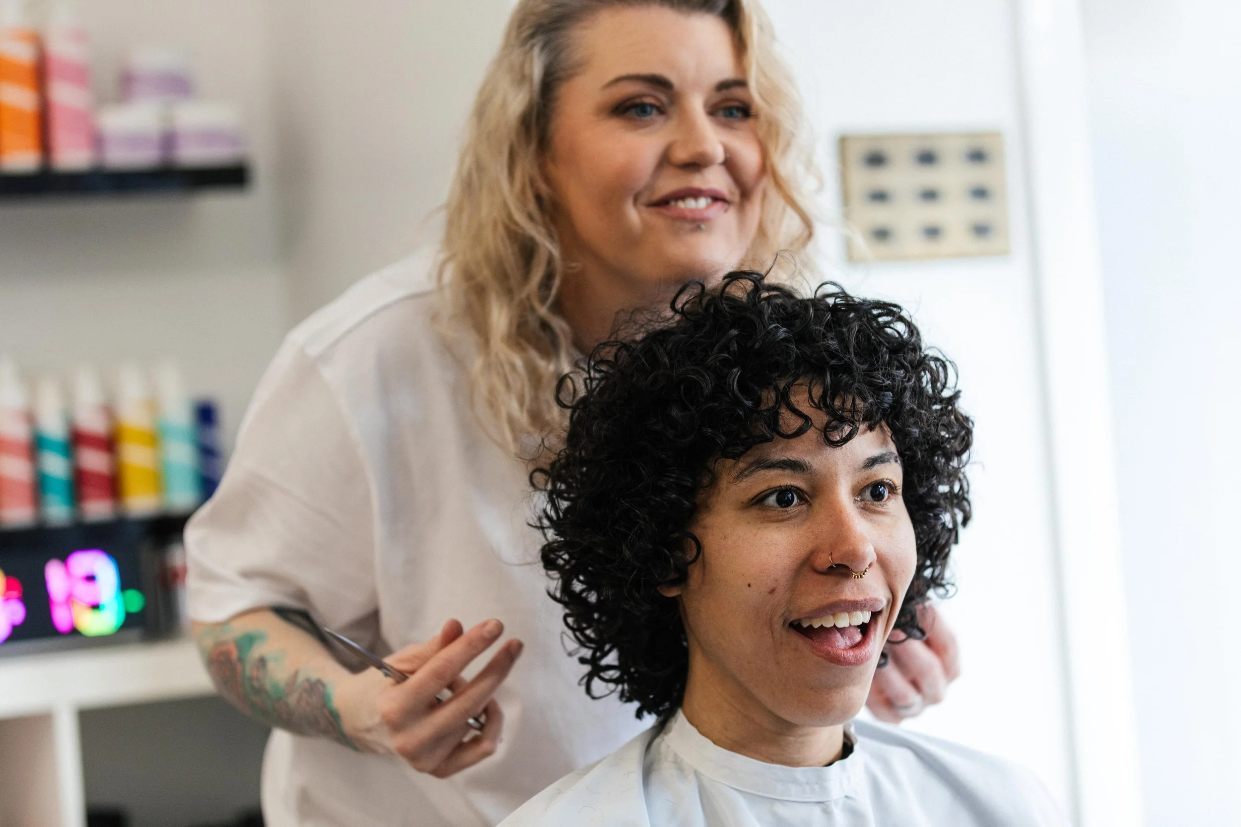 A woman with blonde hair and tattoos on her arm is smiling while a hairstylist with curly hair cuts her hair in a salon.