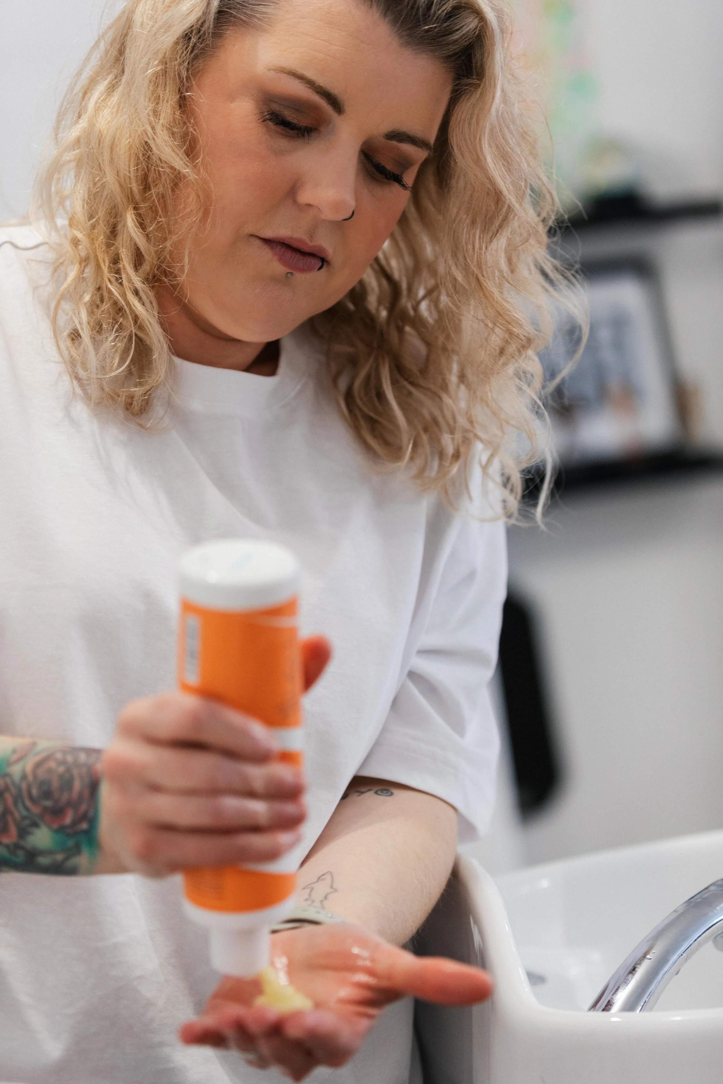A woman with blonde, curly hair applying lotion or cream to her hand over a sink.