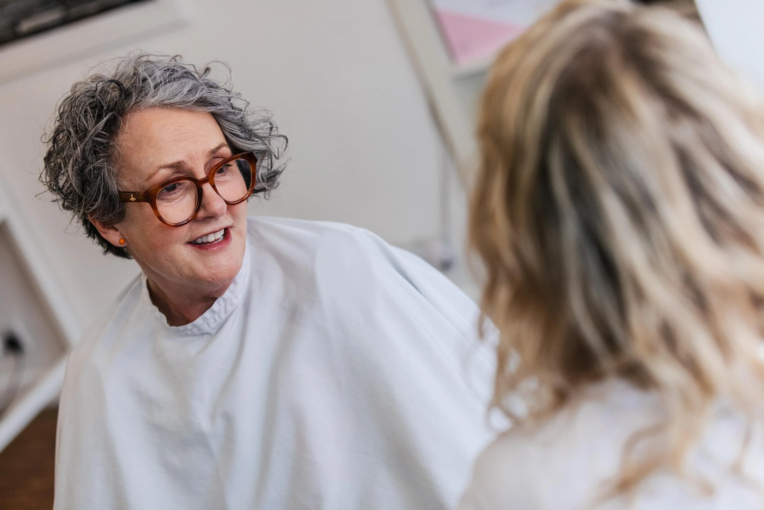 A woman with curly gray hair and glasses, wearing a white gown, is talking to another woman with blonde hair, in a salon or similar setting.