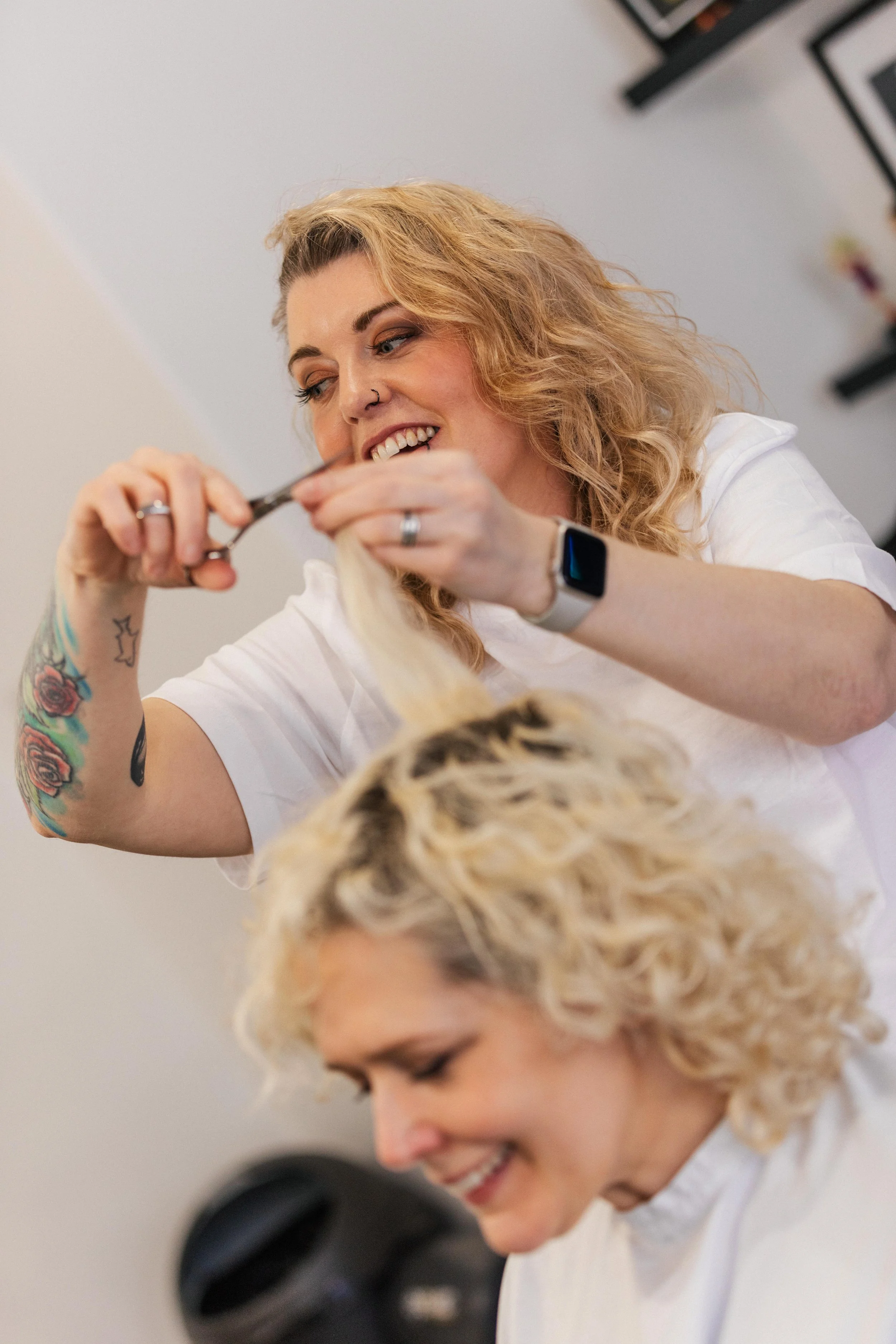 A hairstylist with blonde, curly hair and tattoos on her arm is cutting a woman's blonde, curly hair with scissors. Both women are smiling.