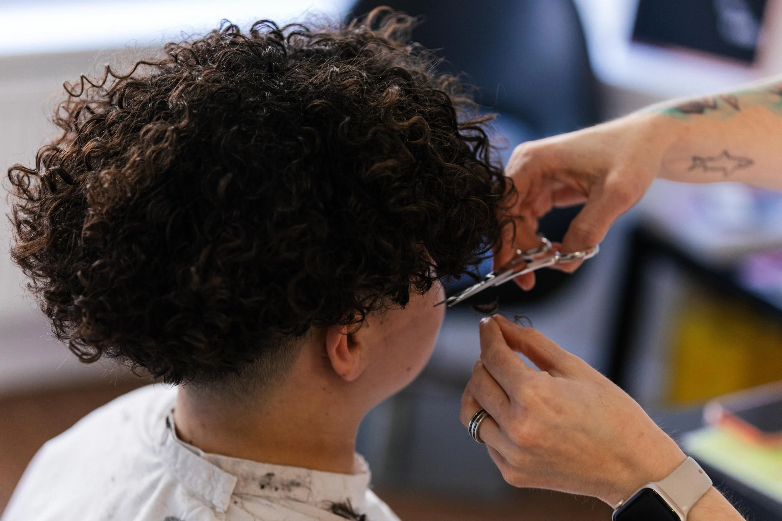 Person with curly hair getting a haircut at a salon, with a stylist using scissors.