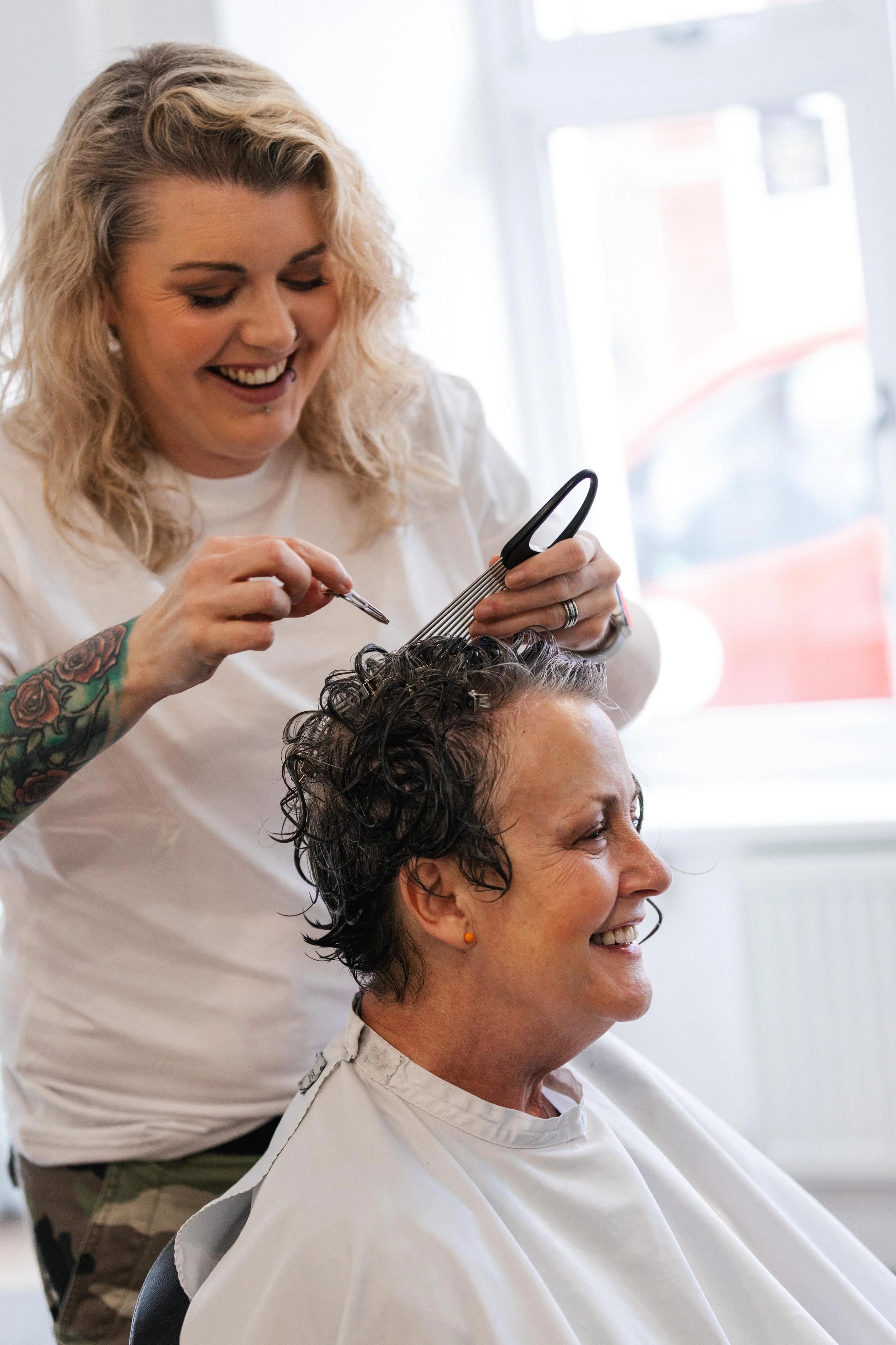 A woman with short curly dark hair sitting in a salon chair, smiling as a hairstylist with blonde curly hair and tattoos on her arm cuts and styles her hair.