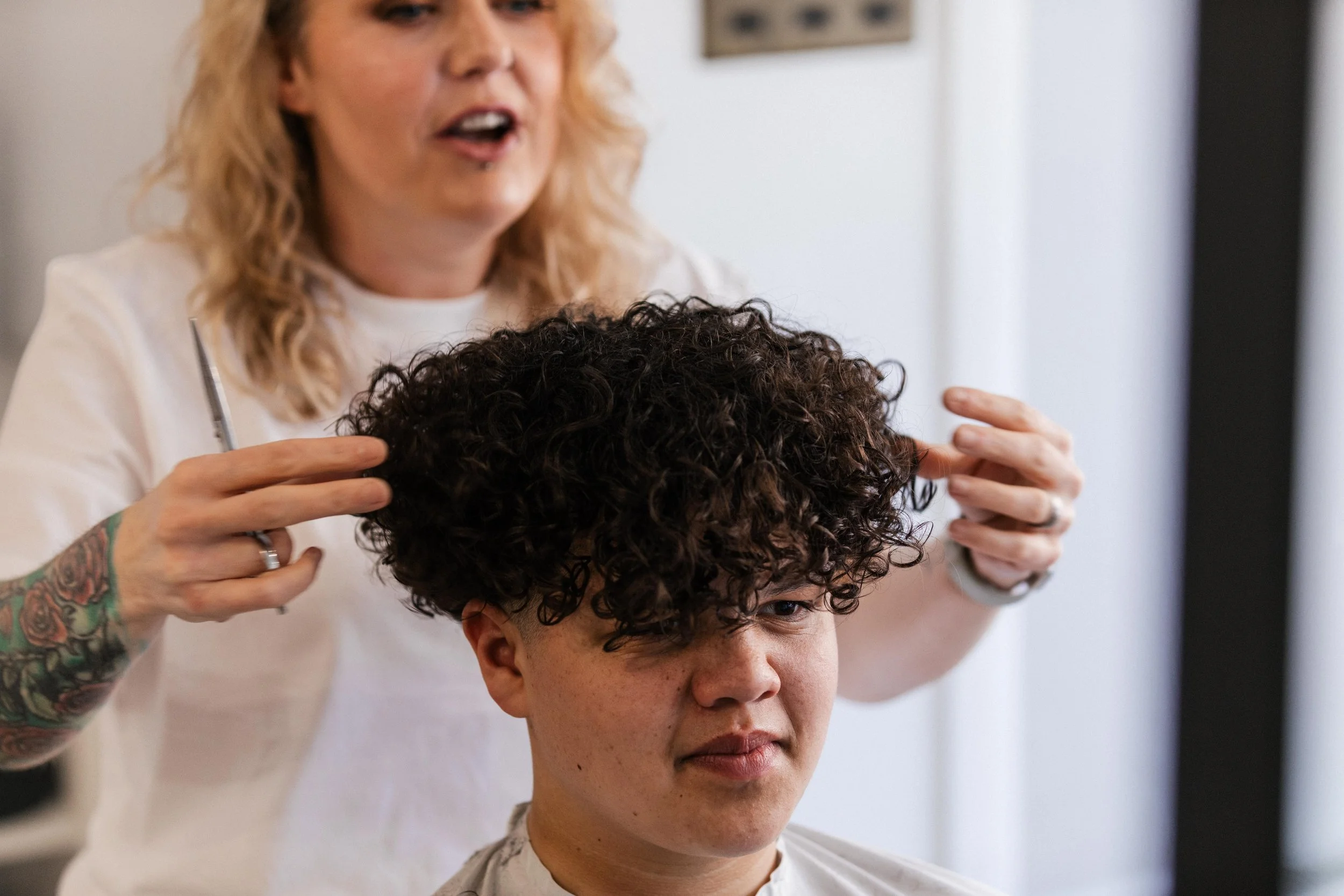 A woman with blonde hair and tattoos on her arm is styling a young man's curly hair in a salon.