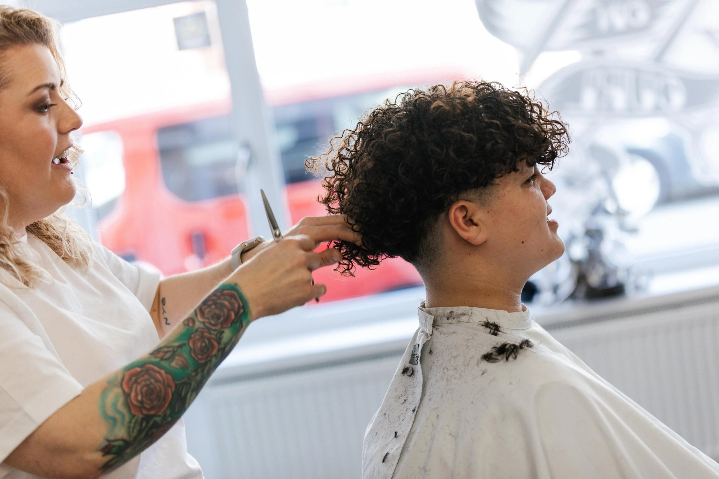 A woman with tattoos on her arm is cutting a person's curly hair in a salon, with a window showing a red vehicle outside.