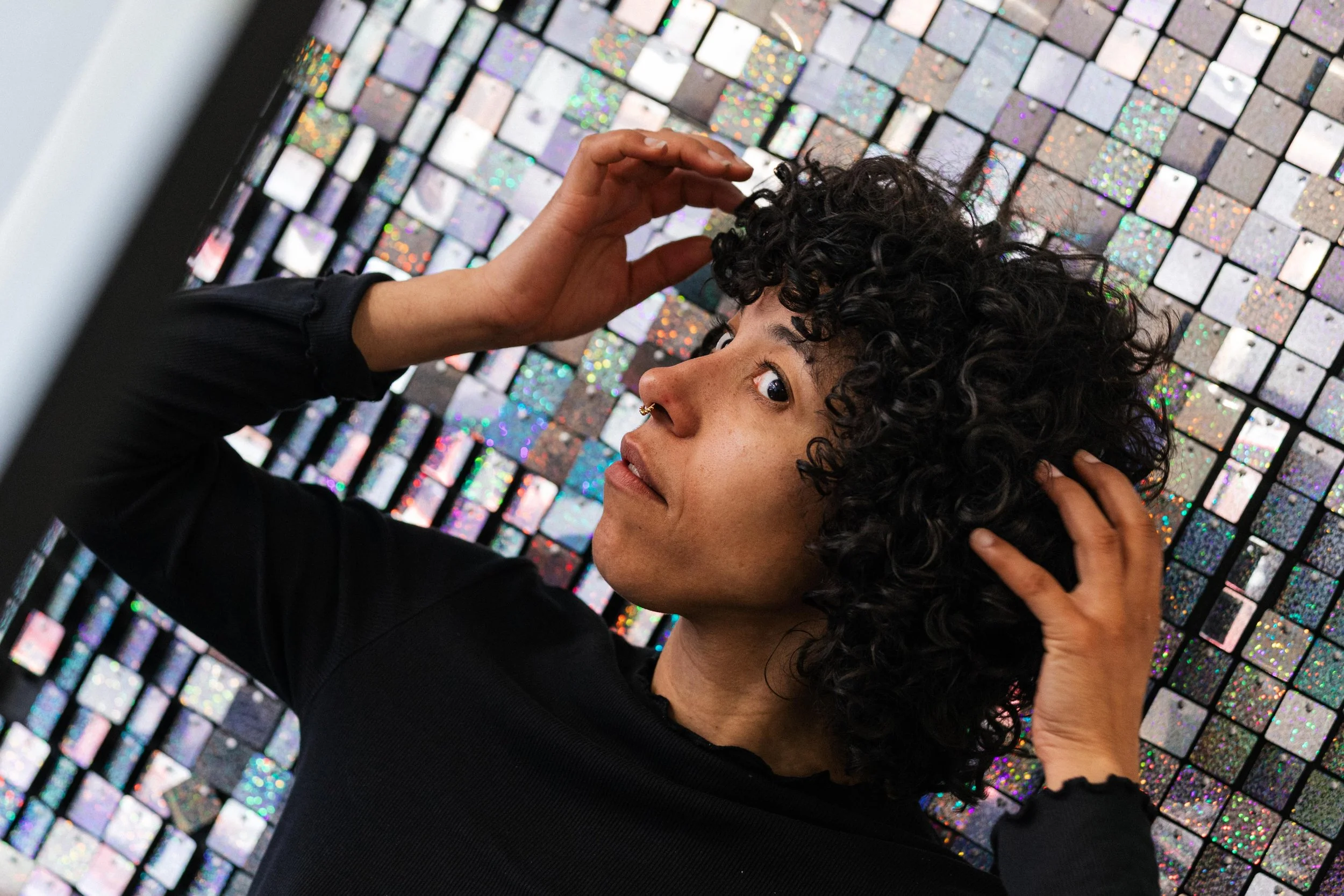 Woman with curly hair and a septum piercing posing in front of a reflective, mosaic wall made of small holographic tiles.