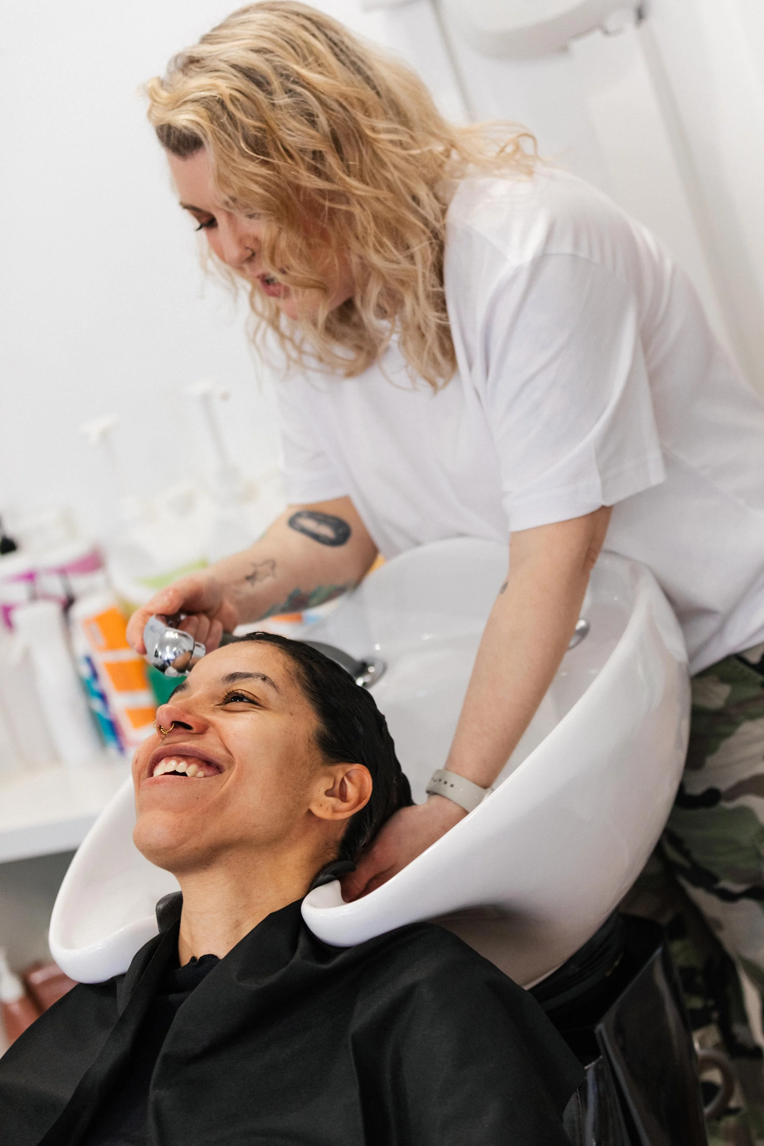 A person getting their hair washed at a salon, smiling, with a stylist washing their hair over a white sink.