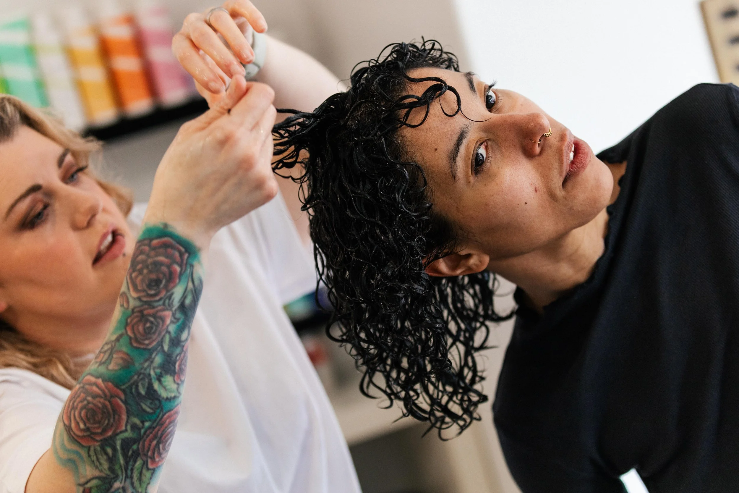 A person with curly black hair getting a haircut from a stylist with tattoos on their arm.