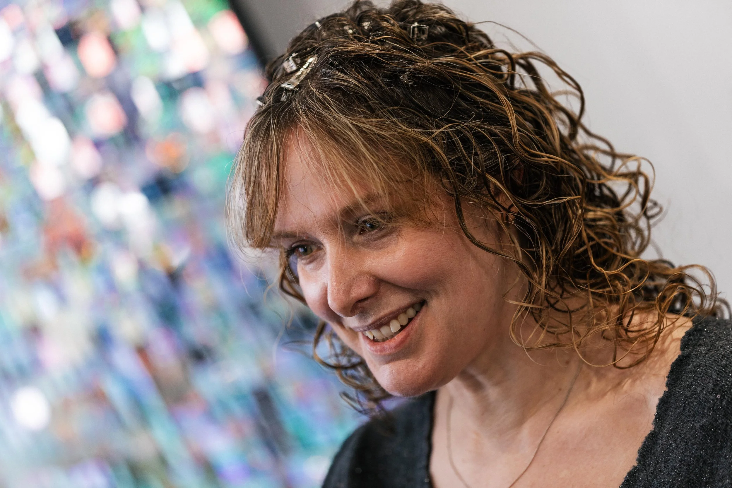 A woman with curly hair smiling and looking down, with a colorful, blurred background.