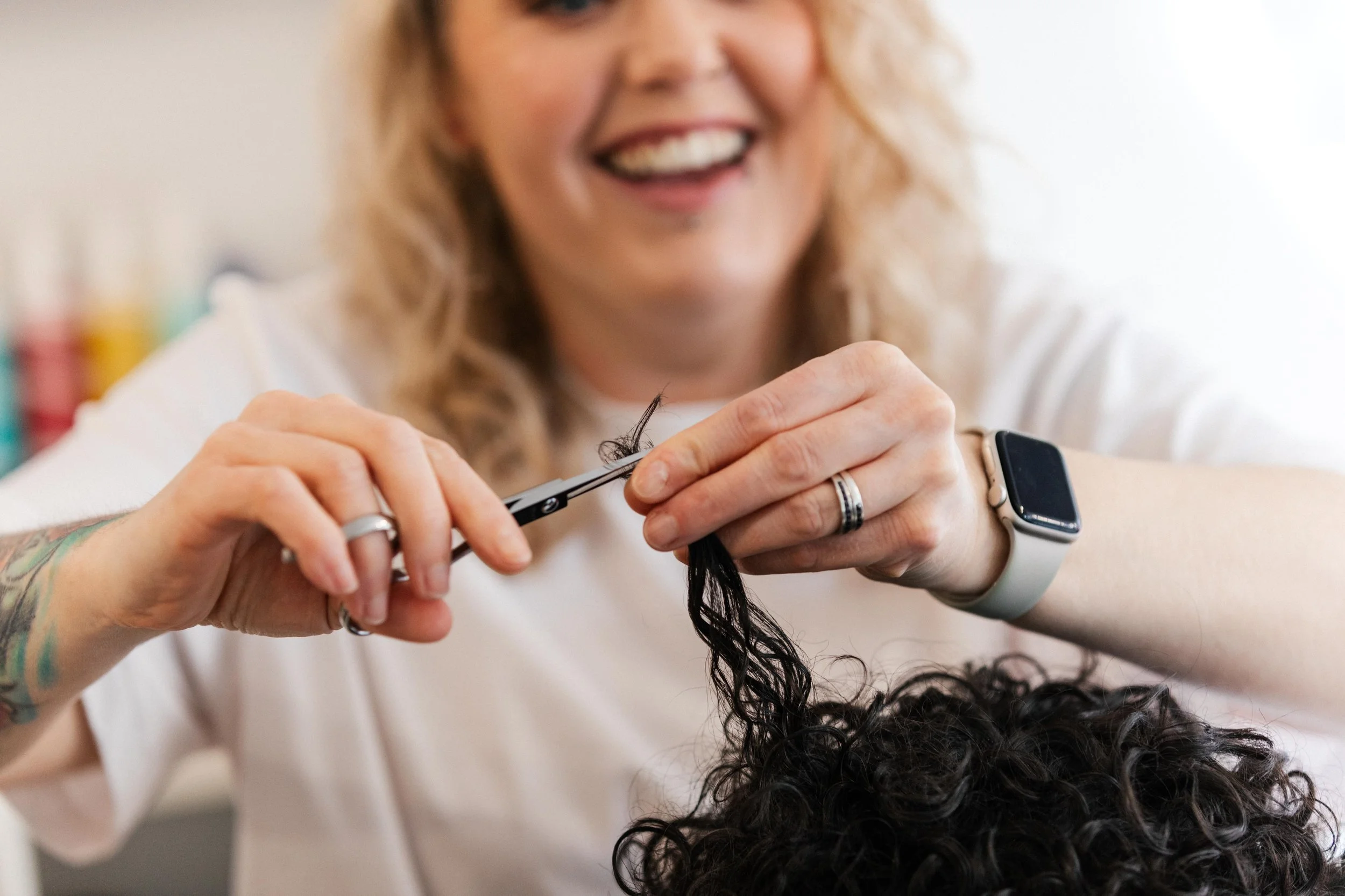 A woman smiling while cutting a person's curly black hair with scissors in a salon.