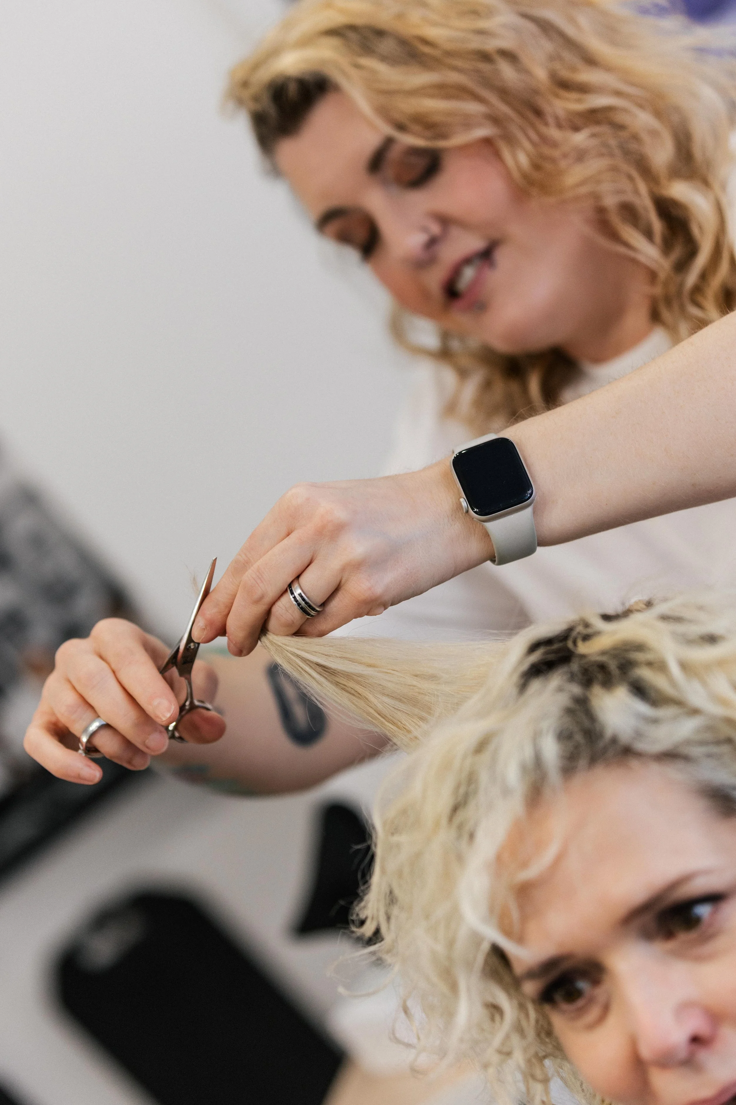 A hairstylist with a smartwatch and rings is cutting a woman's blonde hair with scissors in a salon. The woman is looking at her reflection.