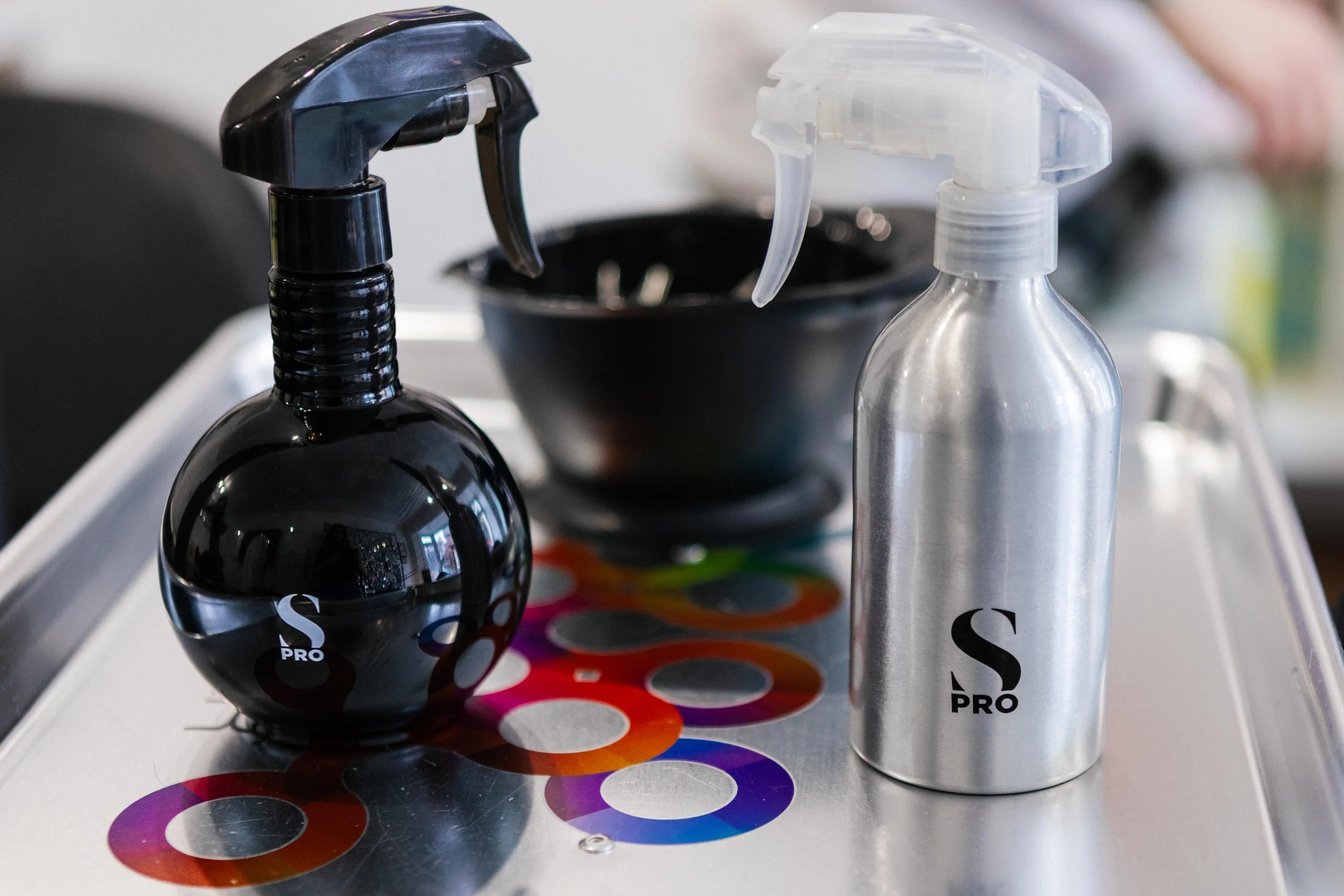 A black spray bottle and a silver spray bottle on a table with color swatches and a black bowl in the background.