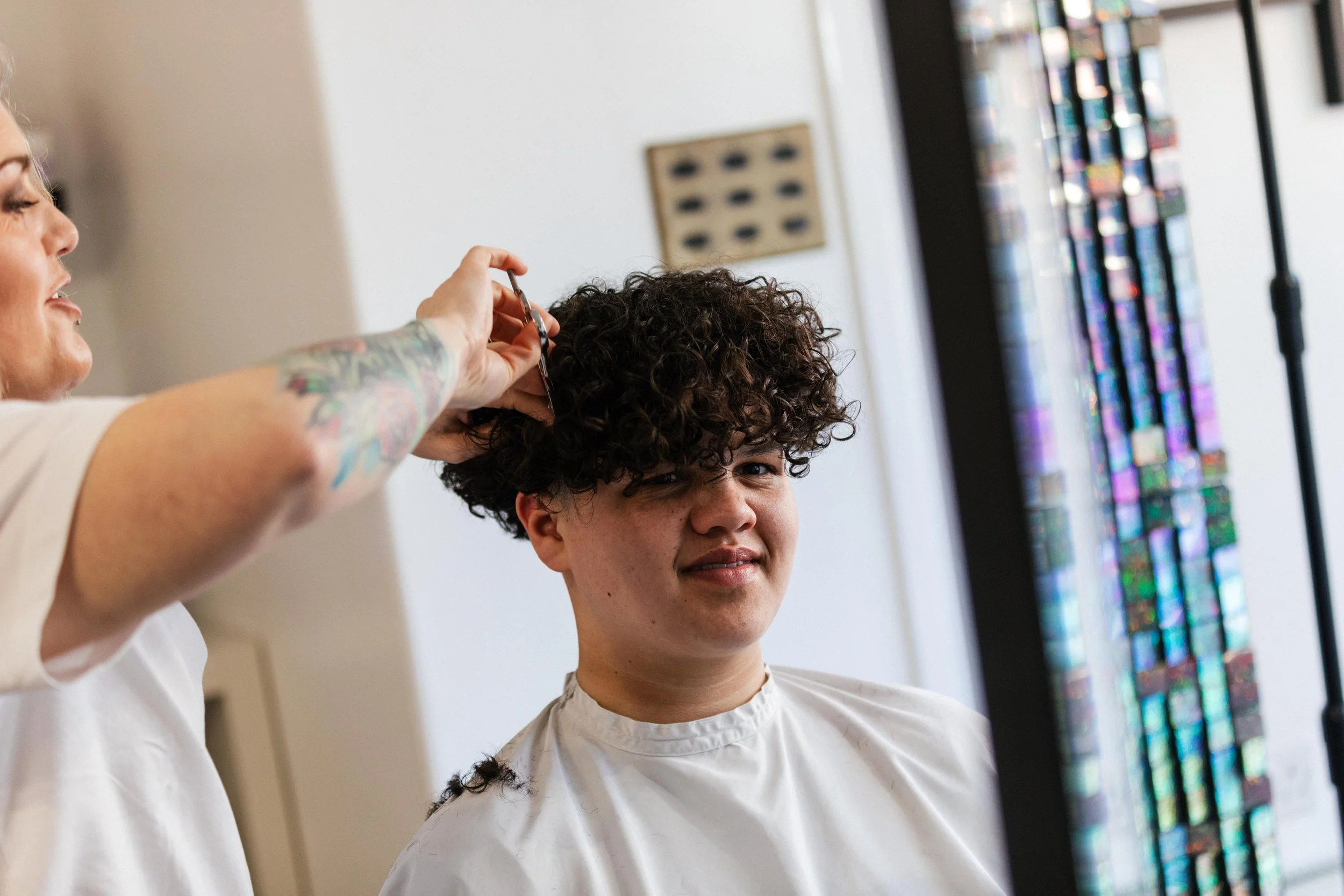 A hairstylist with colorful tattoos on her arm is cutting a young man's curly hair in a salon. The young man is looking in a mirror, making a slight smile.