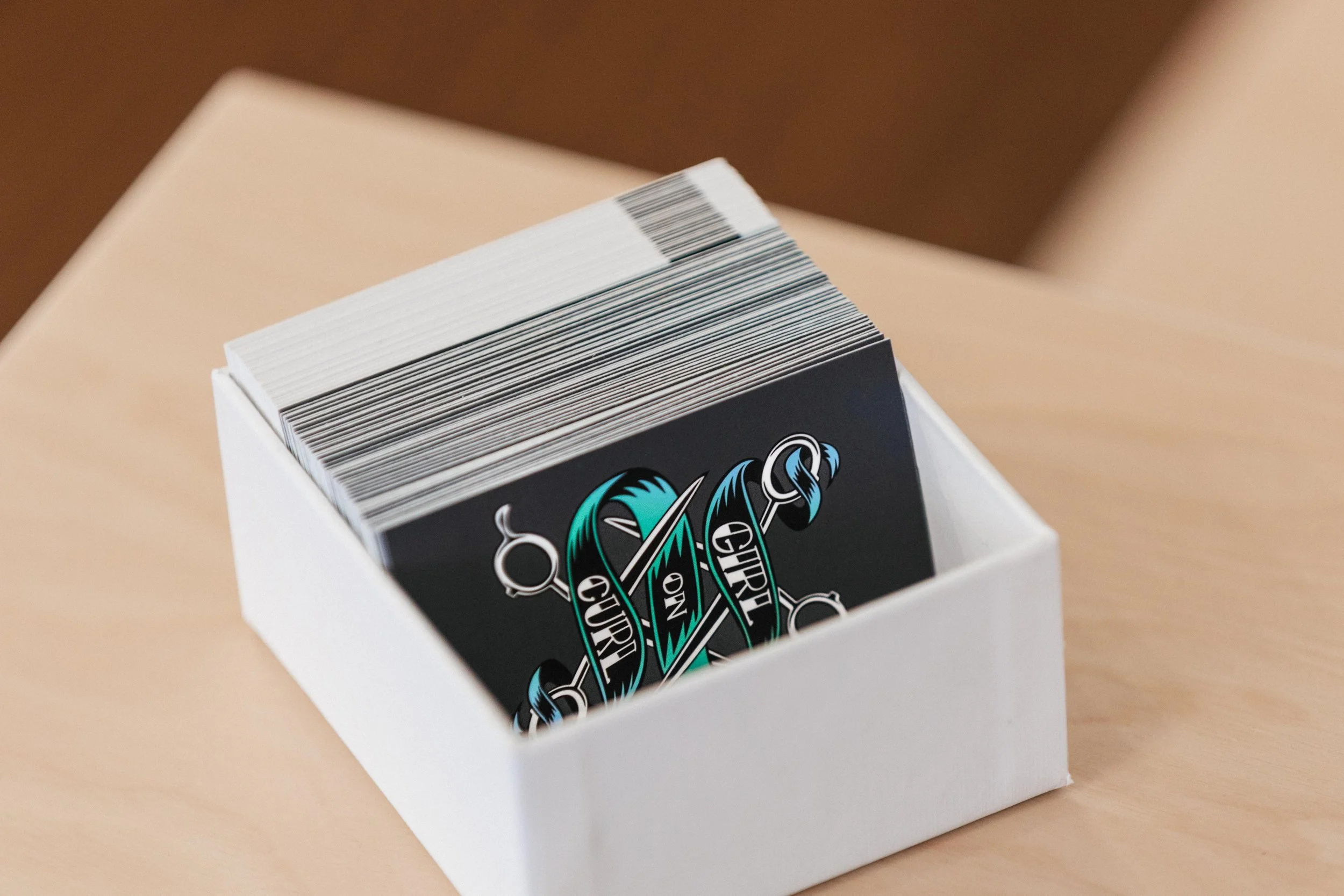 Stack of black cards with a tattoo-style design reading "Girl on Girl" in a white cardboard box on a light wooden surface.
