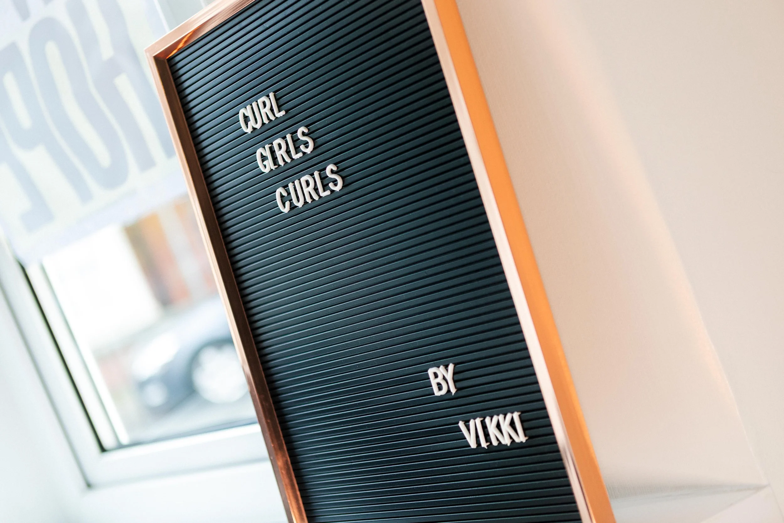Letter board with the words "CURL GIRLS CURLS BY VEKKI" inside a room with a window in the background.