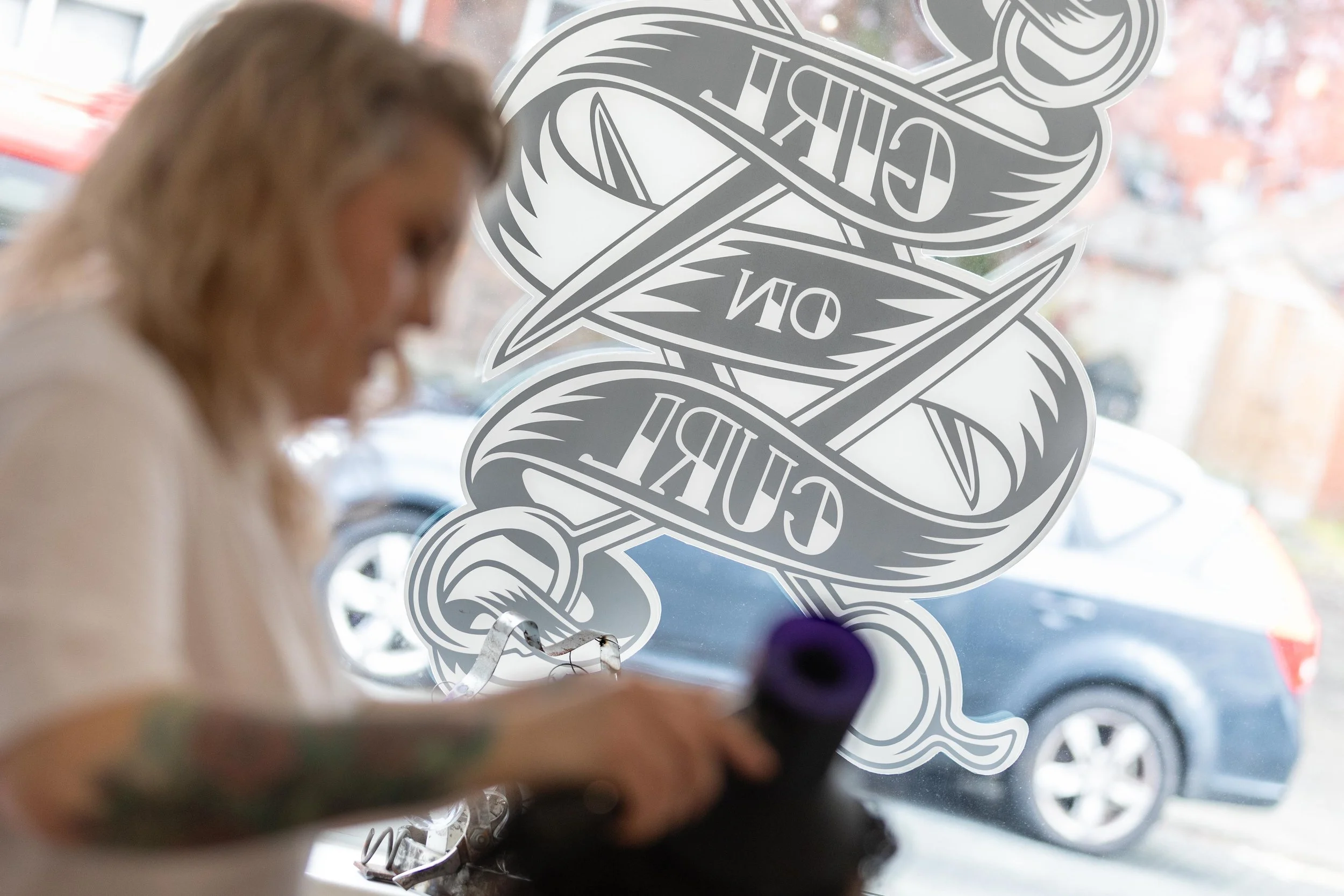 A woman is working on a tattoo in a studio, seen through a window with a large design that says 'GURT ON EDGE' on it.