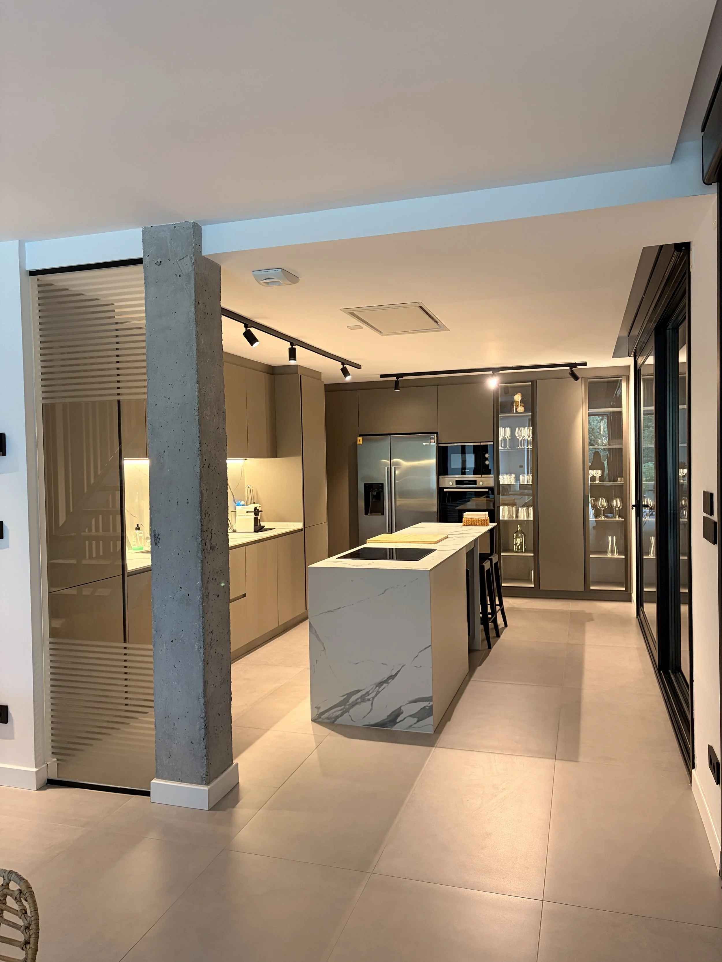 Modern kitchen with beige cabinets, stainless steel appliances, a white marble island, and glass-fronted cabinets displaying glassware.