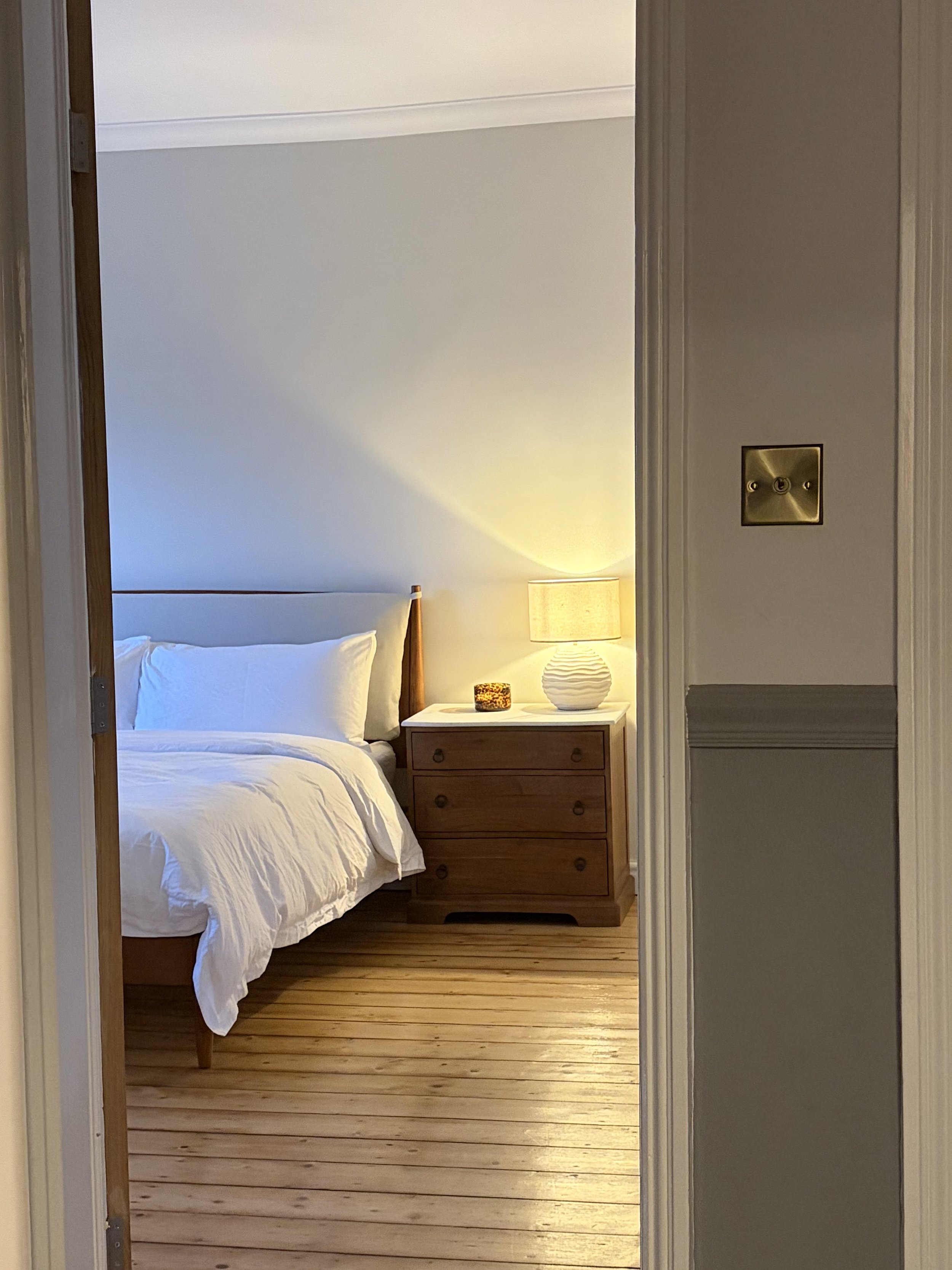 A bedroom with a bed, a wooden nightstand with a lamp and decorative item, and hardwood floors, viewed through a doorway.