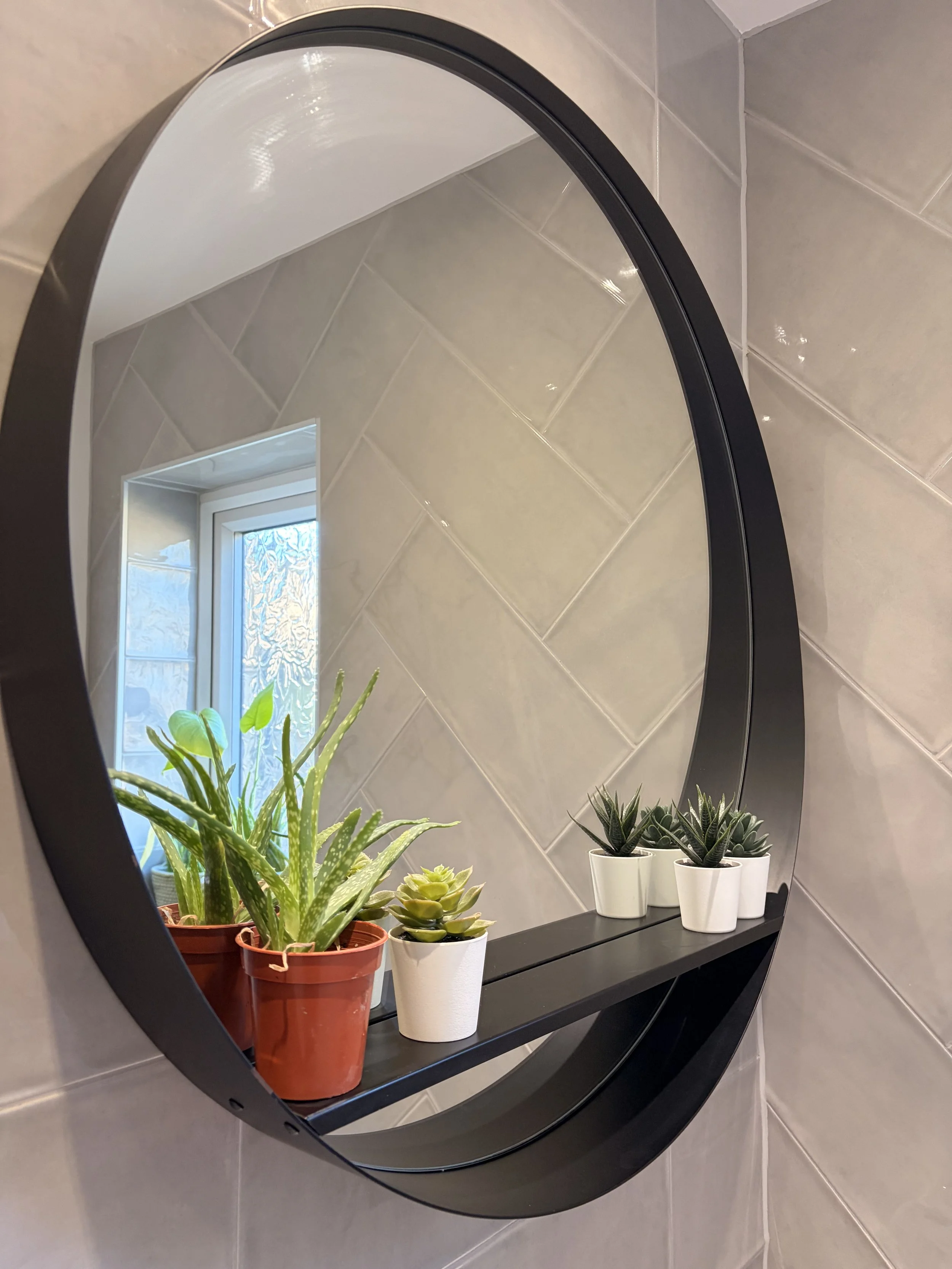 A round black-framed mirror mounted on a tiled wall, reflecting a window with frosted glass and potted plants on a black shelf.