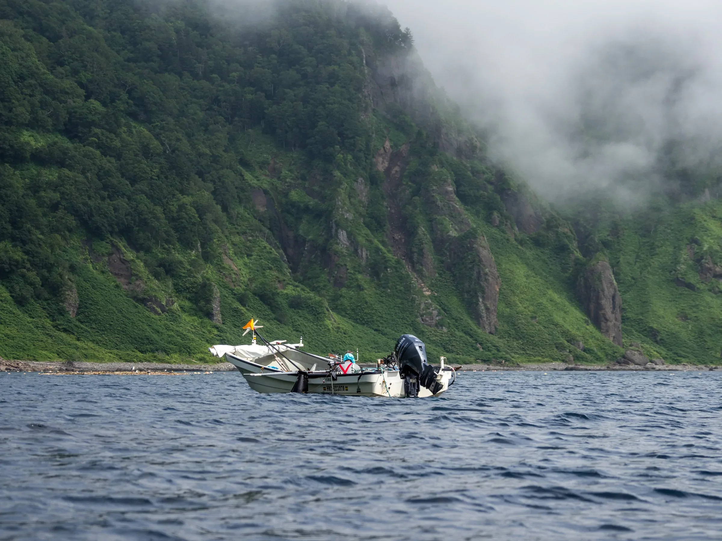 A boat with an outboard motor floating on a body of water near a green, forested mountain with mist or fog around it.