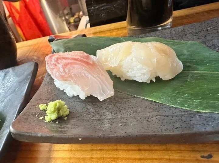 Two pieces of sushi, one with a pale pink fish and the other with white fish, served on a green leaf on a dark rectangular plate, with a small mound of wasabi nearby.