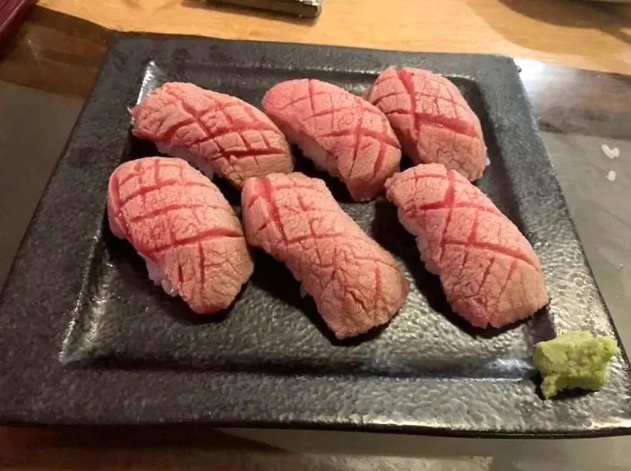 Seven pieces of sushi with pink, fatty tuna on a black rectangular plate, garnished with a small amount of wasabi on the side.