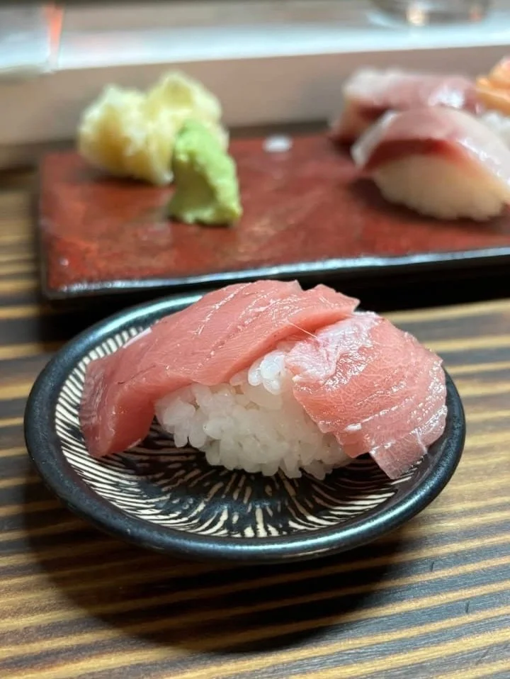 Close-up of a piece of tuna nigiri sushi with white rice underneath, placed on a black decorative plate on a wooden table. In the background, a red rectangular plate holds wasabi and additional sushi pieces.