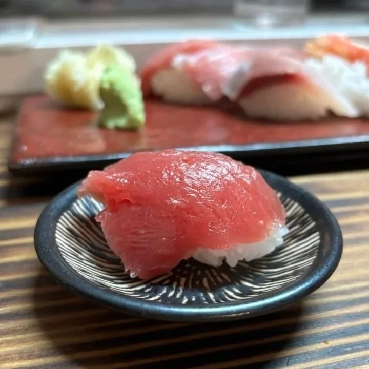 A close-up of a piece of tuna nigiri sushi on a black ceramic plate, with wasabi and more sushi in the background on a red plate.