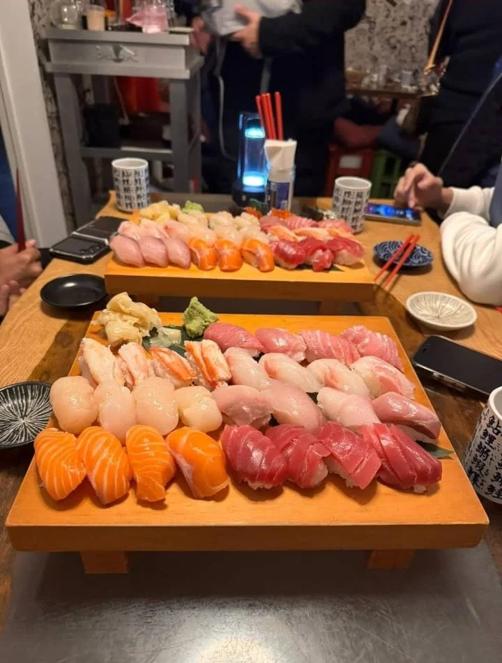 Two wooden trays filled with assorted sushi, including salmon, tuna, shrimp, and other fish, served with wasabi and ginger, on a dining table with several cups and bowls, surrounded by people at a restaurant.