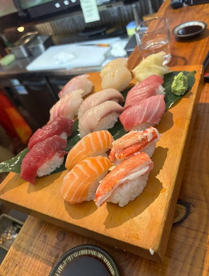 Assorted sushi on a wooden serving board with wasabi, ginger, and soy sauce, with a glass of water and a small dish of soy sauce in the background.