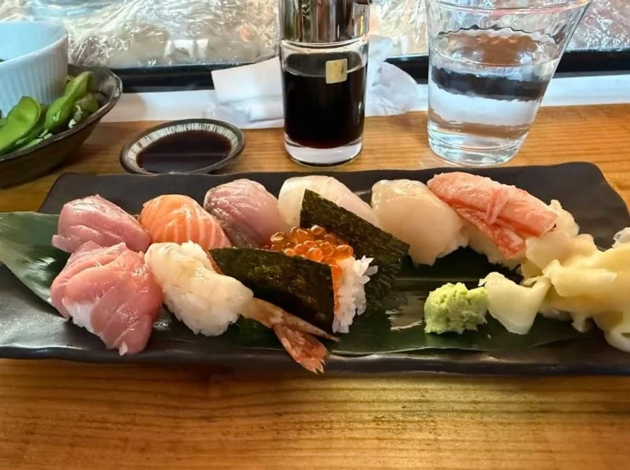 Assorted sushi on a black rectangular plate, including raw fish, shrimp, fish roe, and pickled ginger, with wasabi on the side. In the background, there is a glass of water, soy sauce, a small dish of soy sauce, a bottle of soy sauce, a bowl of green vegetables, and a glass of water on a wooden table.