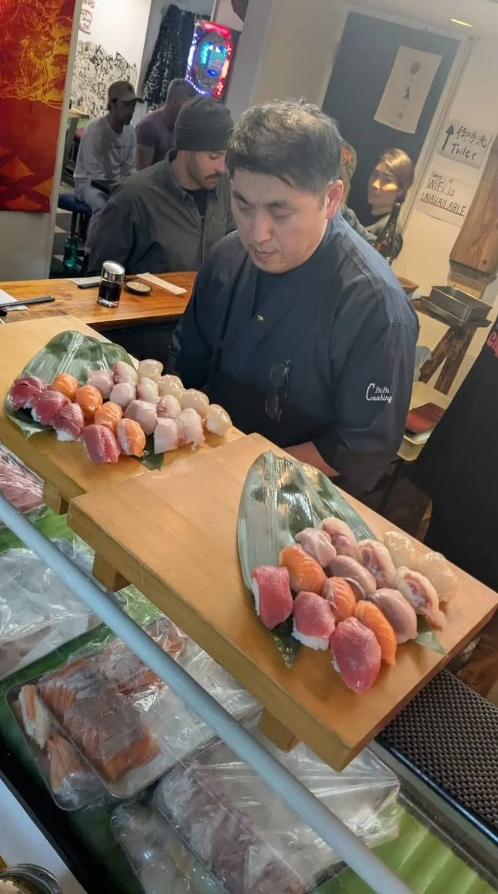 A sushi chef preparing an assortment of sushi and sashimi on two wooden boards in a restaurant, with customers seated in the background.