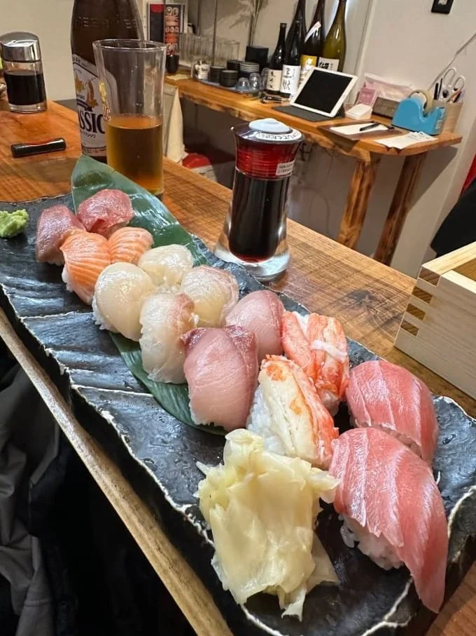 A long black serving plate with assorted sushi pieces, wasabi, and pickled ginger on a wooden table. Two glasses, one filled with beer and the other with soy sauce, are beside the plate. The background shows a bar area with bottles, a tablet, and various bar tools.