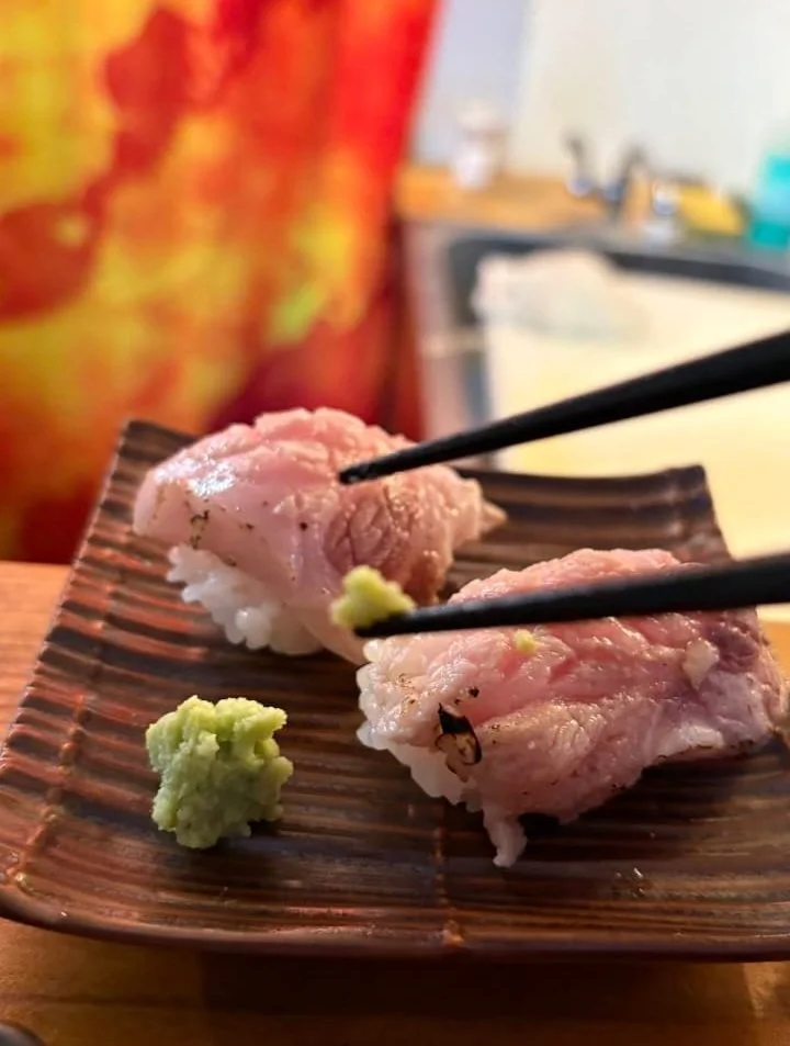 Close-up of two pieces of sushi with wasabi on a ceramic plate, chopsticks holding one piece, kitchen in the background.