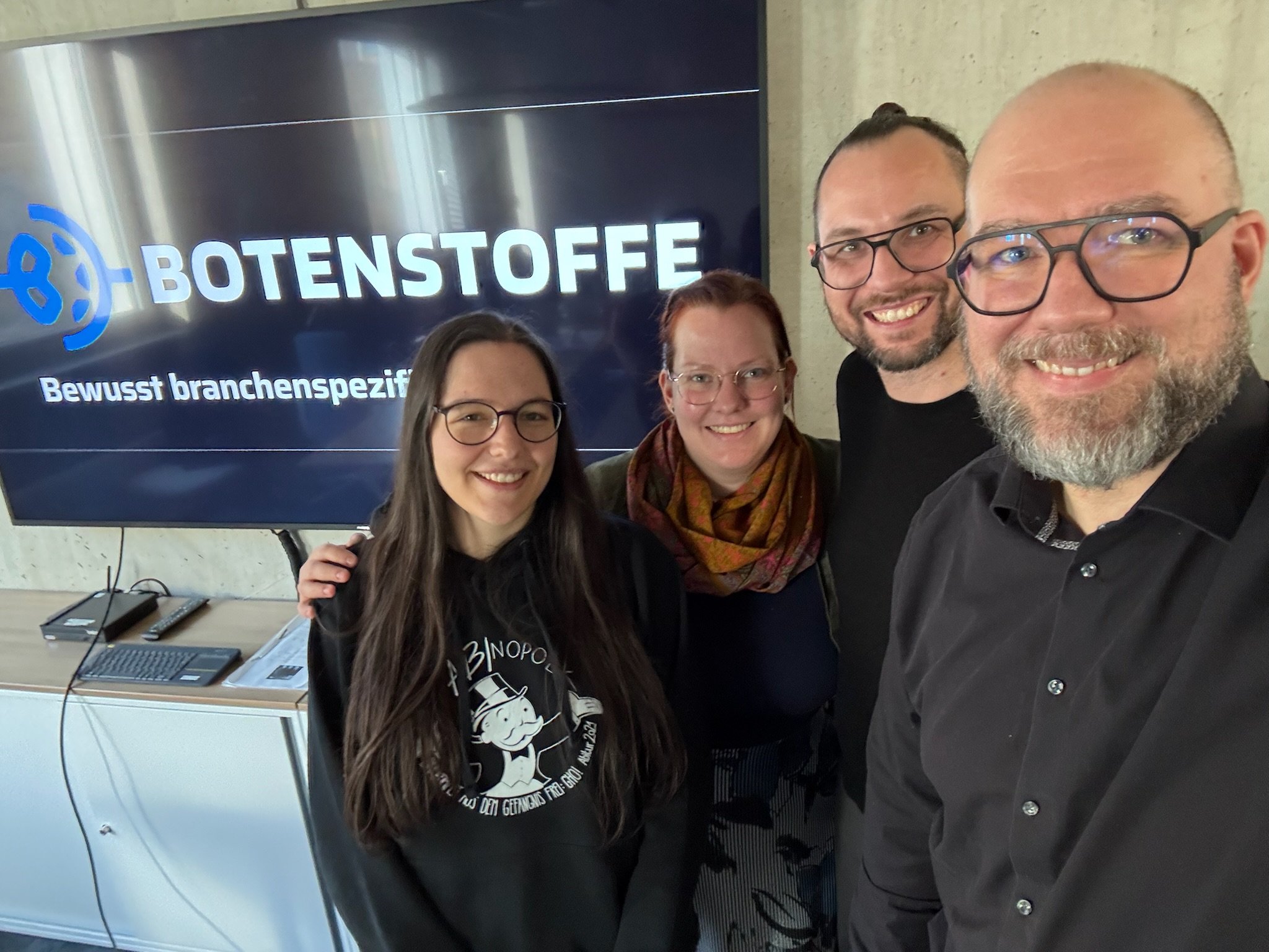 Vier Personen nehmen ein Selfie vor einem Bildschirm mit dem Text 'BOTENSTOFFE Bewusst branchenspezifisch' auf.