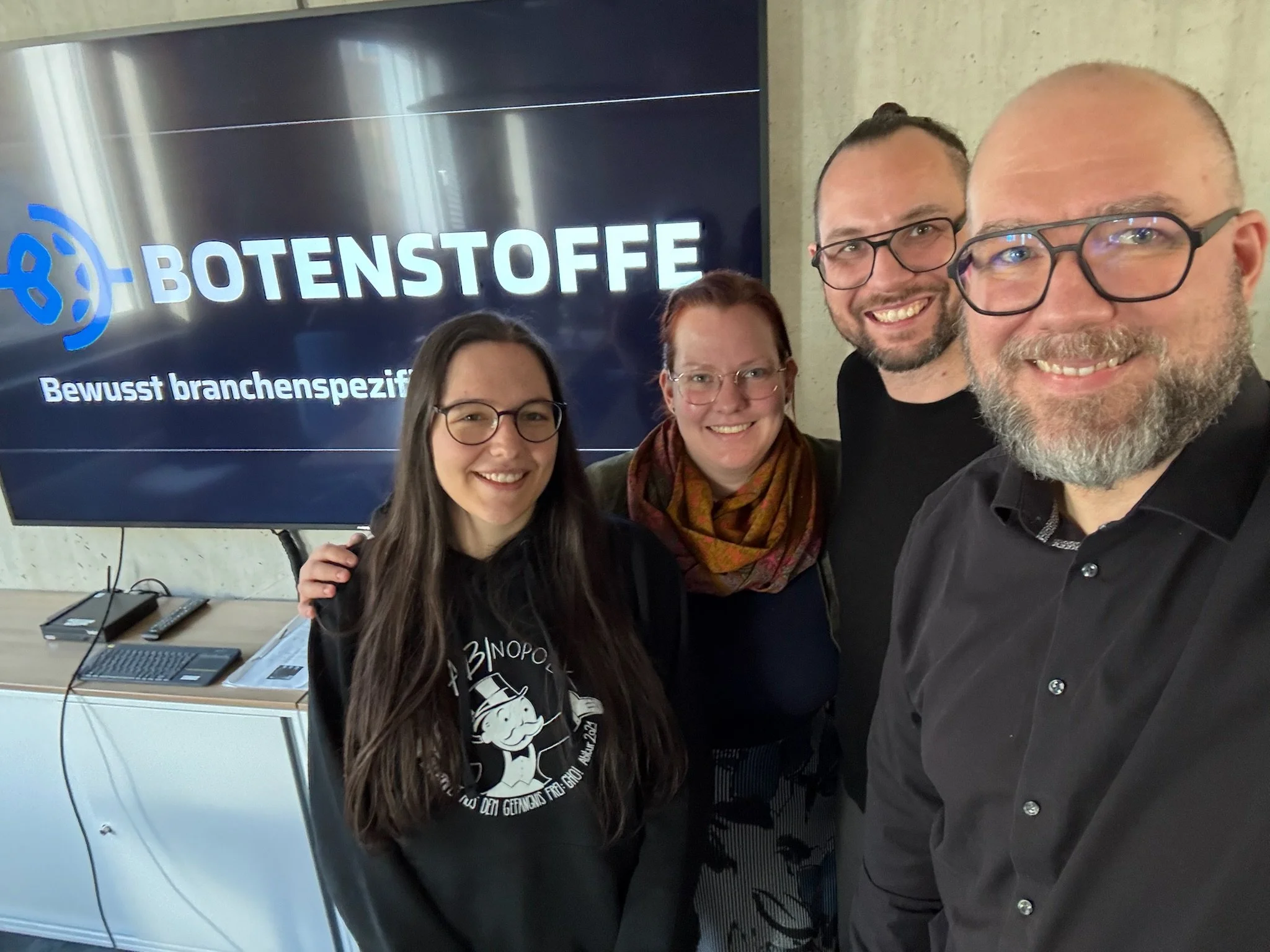 Vier Personen nehmen ein Selfie vor einem Bildschirm mit dem Text 'BOTENSTOFFE Bewusst branchenspezifisch' auf. Hannah Rosenbaum, Jessica Hertenstein, Karsten Hellinger, Rüdiger Wolf-Sebottendorff.
