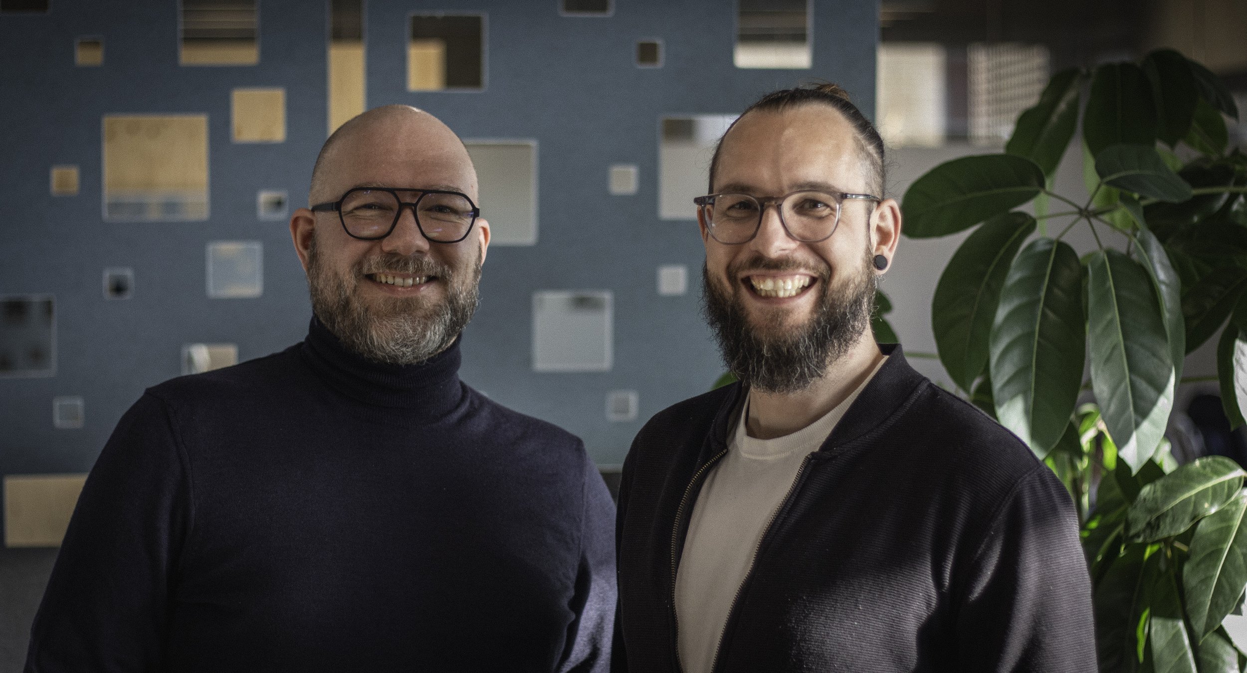 Zwei lachende Männer mit Brillen und Bart in einem modernen Büro, im Hintergrund eine blaue Wand mit quadratischen Ausschnitten und eine große grüne Pflanze.