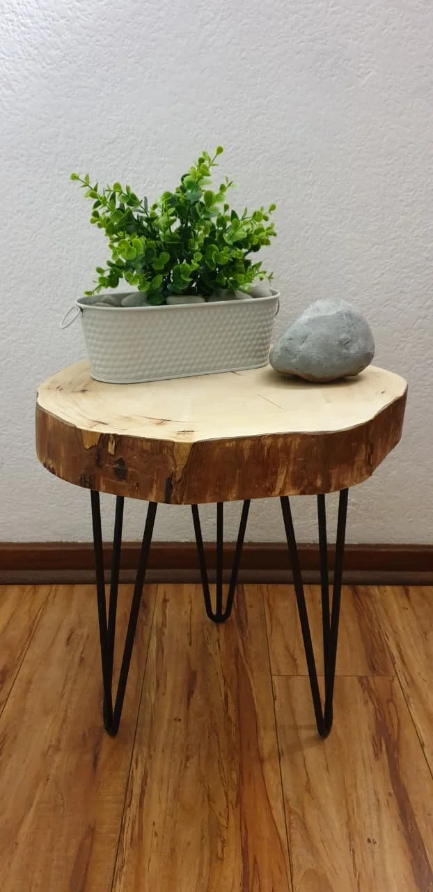 A small wooden side table with black metal hairpin legs, topped with a rectangular white textured planter with a green artificial plant and a smooth gray stone, set against a plain beige wall with wooden floor.