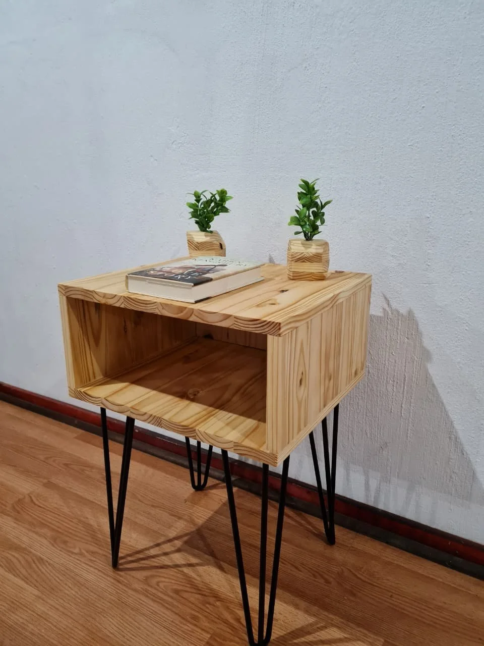 Wooden side table with black hairpin legs, on a hardwood floor, against a textured white wall, decorated with two small potted green plants and a book.
