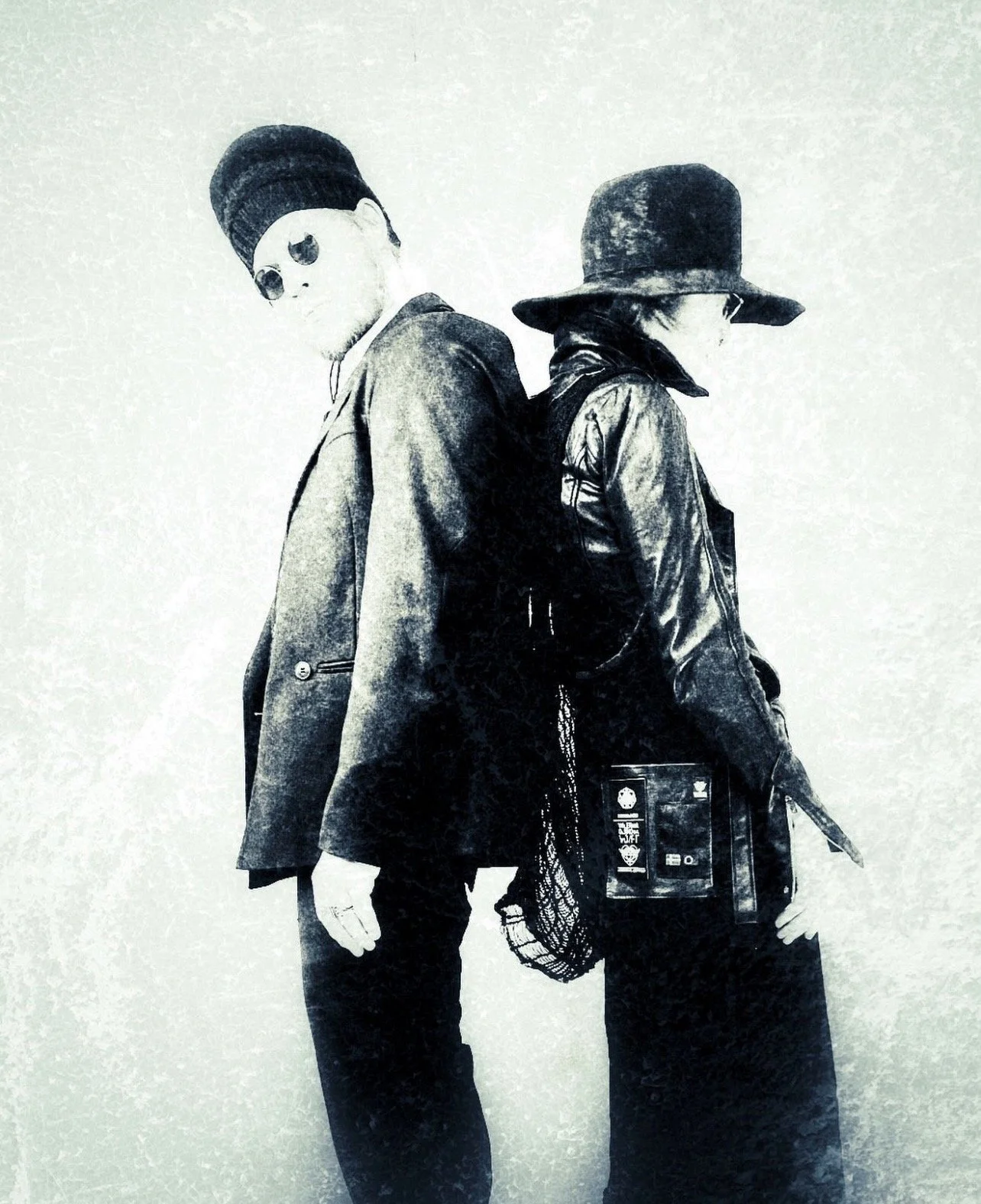 Leon Louis and Valeriya Olkhova - the duo couple standing back-to-back, wearing wide-brimmed hats, circular vintage sunglasses (sygekasse briller) , and dark avantgarde clothing, with an off-white distressed background.
