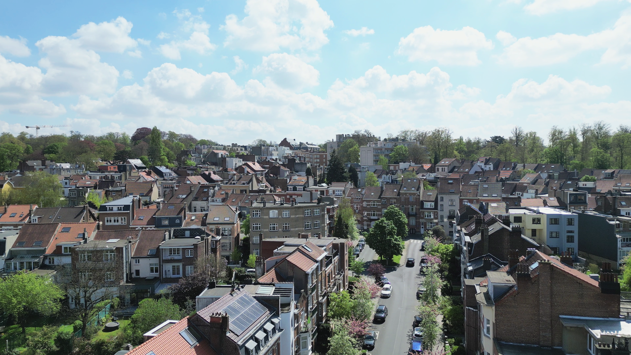 Vue aérienne d'une ville résidentielle avec maisons et bâtiments, entourée d'arbres verts, ciel partiellement nuageux.