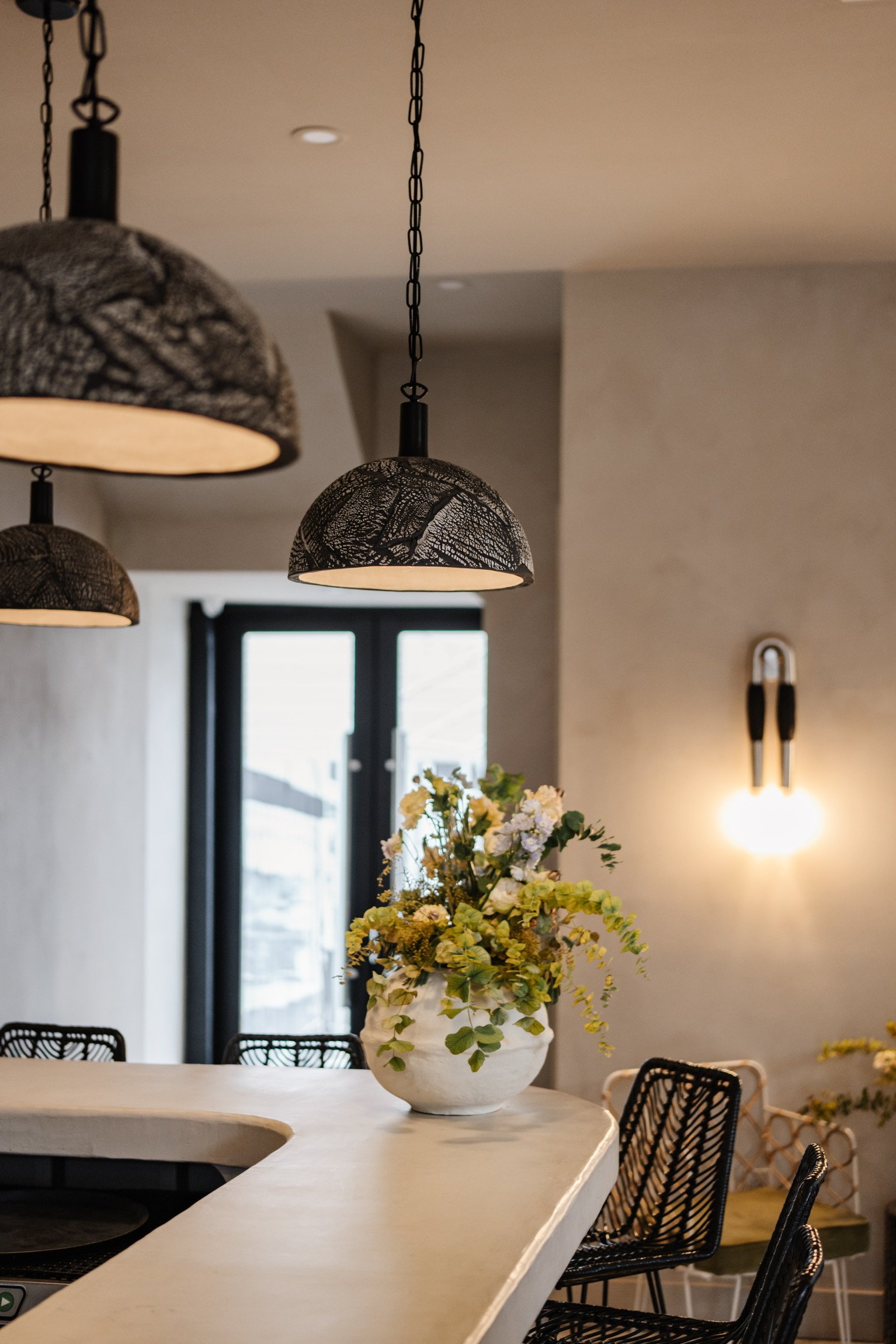 A modern wine bar with pendant lights, a white curved countertop, a large floral arrangement in a white vase, and black wire chairs, with a window in the background.