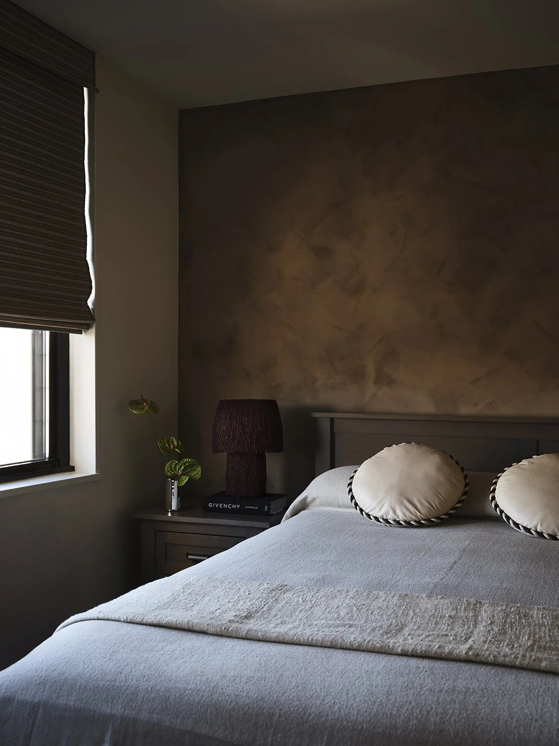 A bedroom with a bed, beige pillows, and a nightstand with a lamp, window with blinds, and a plant.
